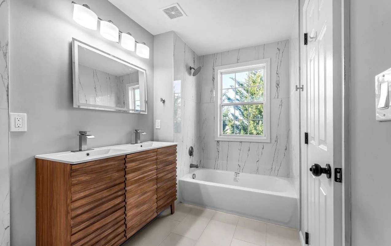 Modern bathroom with double vanity sink, large mirror, wall-mounted lights, bathtub, marble tiles, window showing greenery outside, and closed door.