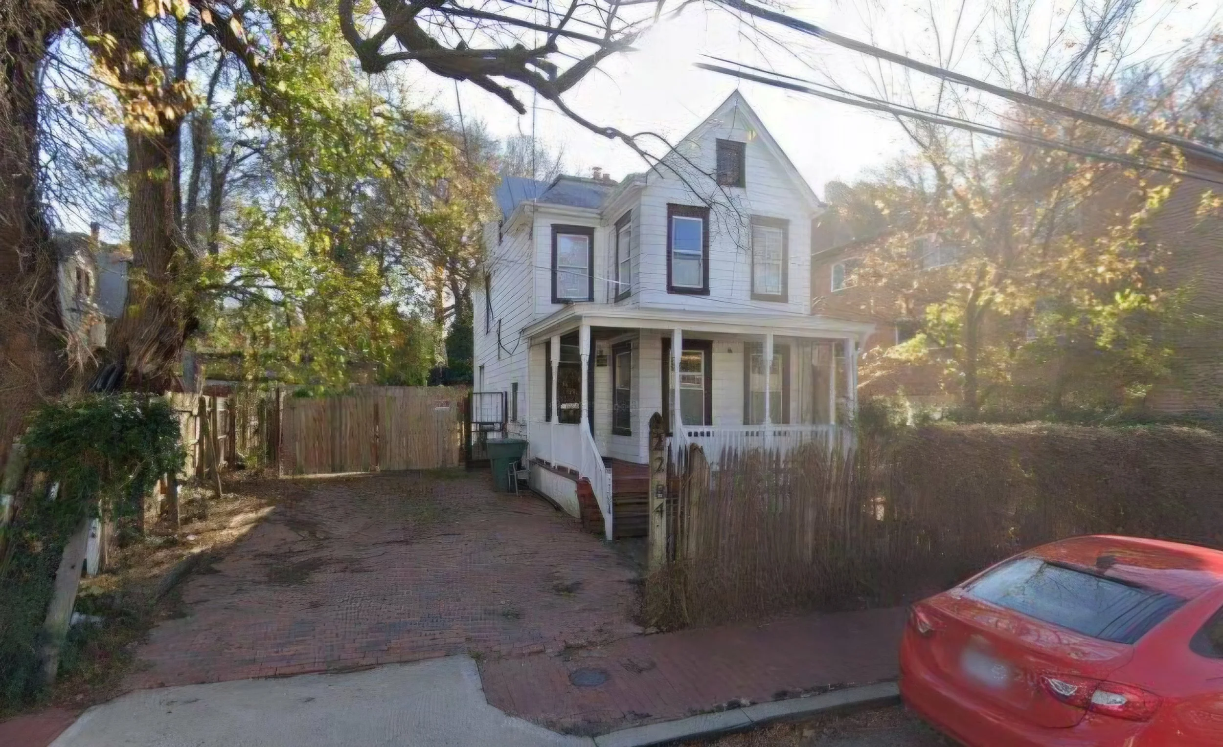 A two-story white house with a front porch, surrounded by trees with autumn leaves, a wooden fence, and a brick-paved driveway with a red car parked at the bottom right corner.