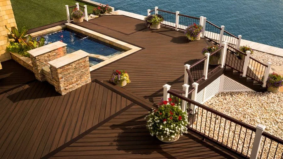 A wooden deck with stairs leading down to a gravel area and a pool, decorated with potted flowers, overlooking the water.
