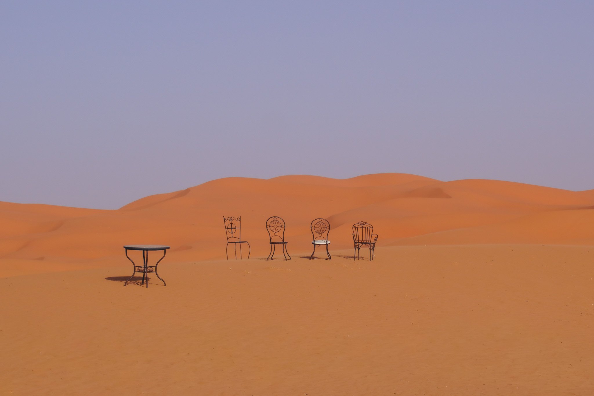 Four decorative metal chairs and one table in a desert landscape on sand dunes under a blue sky.