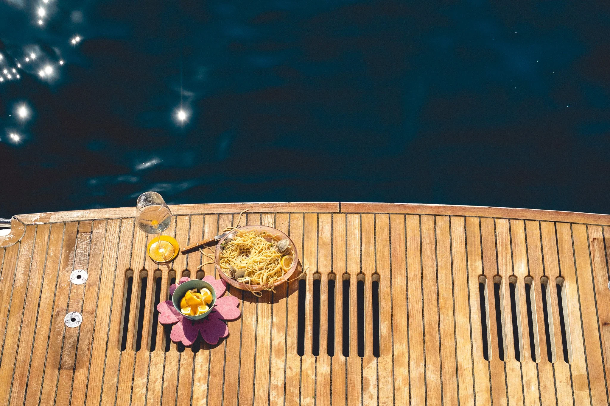 Dinner setup on a wooden dock over water, with a bowl of pasta, a glass of white wine, a small bowl of snacks, and a plate with a flower-shaped placemat.