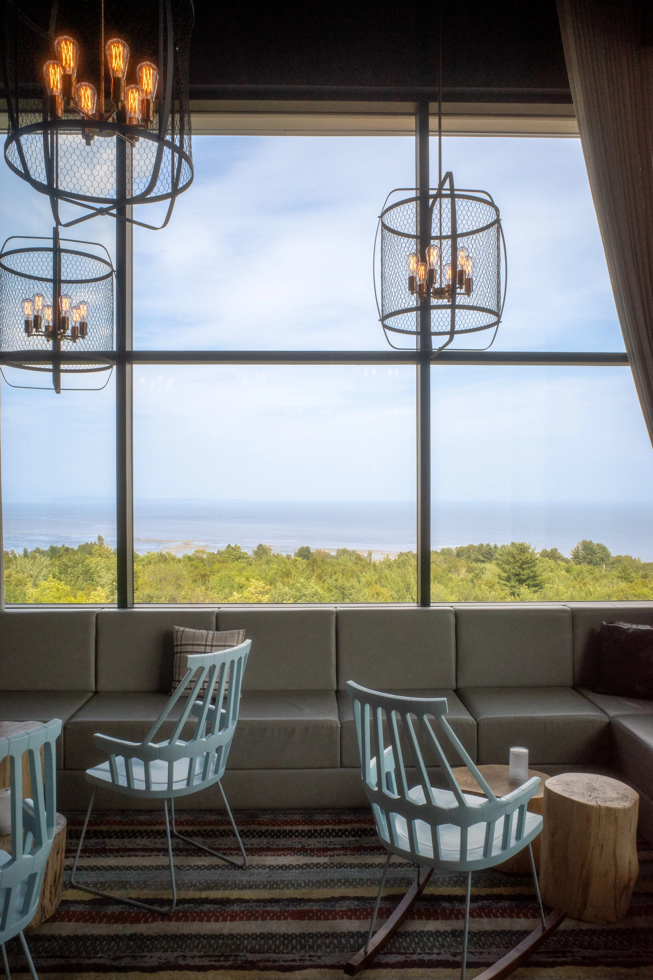 Interior of a room with large window view of a green landscape and ocean, modern hanging chandeliers, light blue chairs, a gray sectional sofa, and wooden side tables.