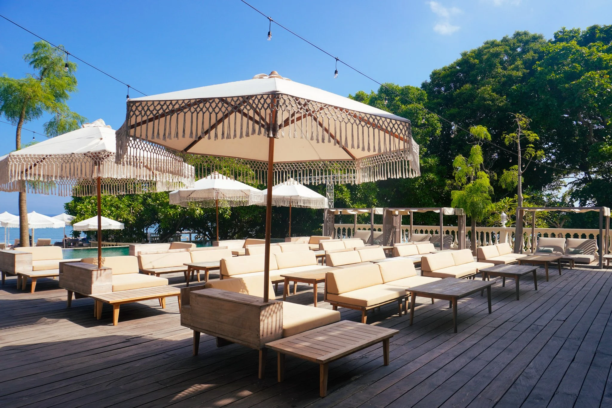 Outdoor lounge area with beige sofas, wooden tables, white umbrellas, and string lights on a wooden deck, surrounded by lush green trees under a blue sky.