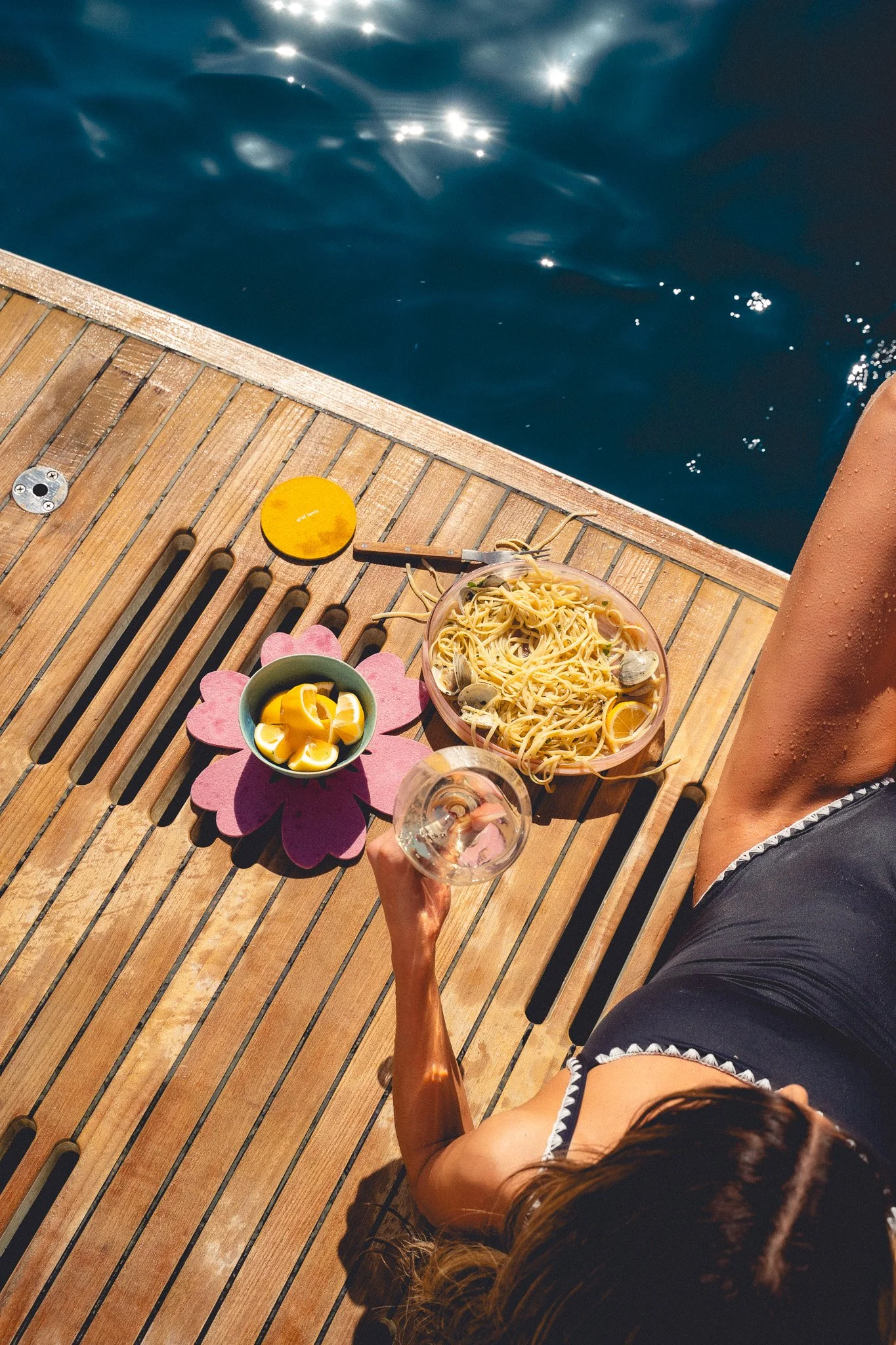 A person sitting on a wooden dock by the water, with a plate of spaghetti, a bowl of lemon wedges, a yellow coaster, and a glass of wine on a pink flower-shaped placemat.