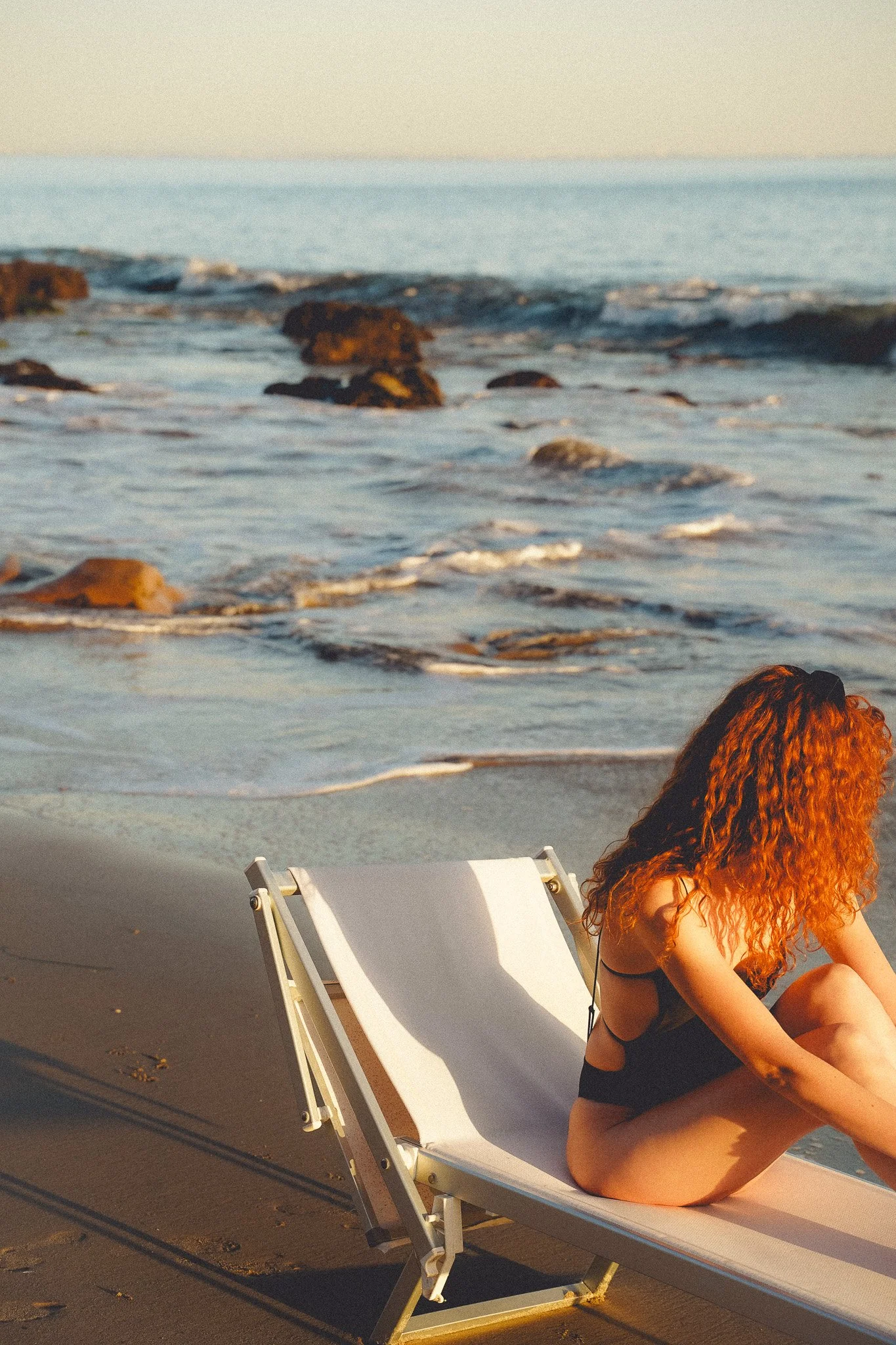 A woman with curly red hair sitting on a beach lounge chair near the ocean, with rocks and waves in the background during sunset.
