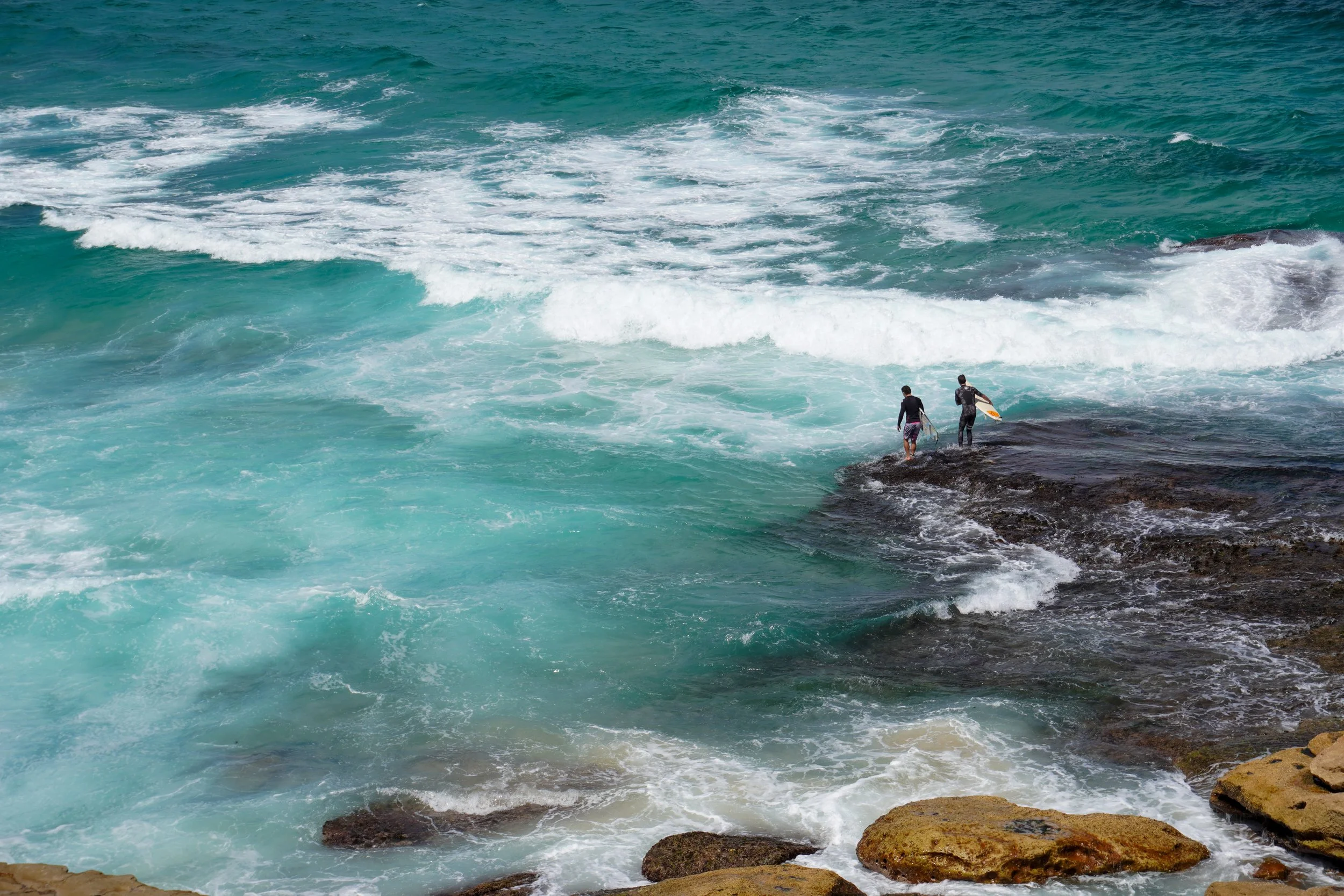 Two surfers in wetsuits walking on rocks near the shoreline with blue-green ocean waves crashing around them.