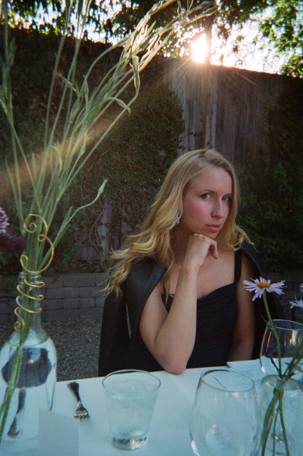 A young woman with long blonde hair sitting at a table outdoors during sunset. She is wearing a black dress and a black jacket, with her chin resting on her hand. The table has a white tablecloth, glasses, forks, and vases with flowers, with the sun setting behind a wooden fence and trees in the background.