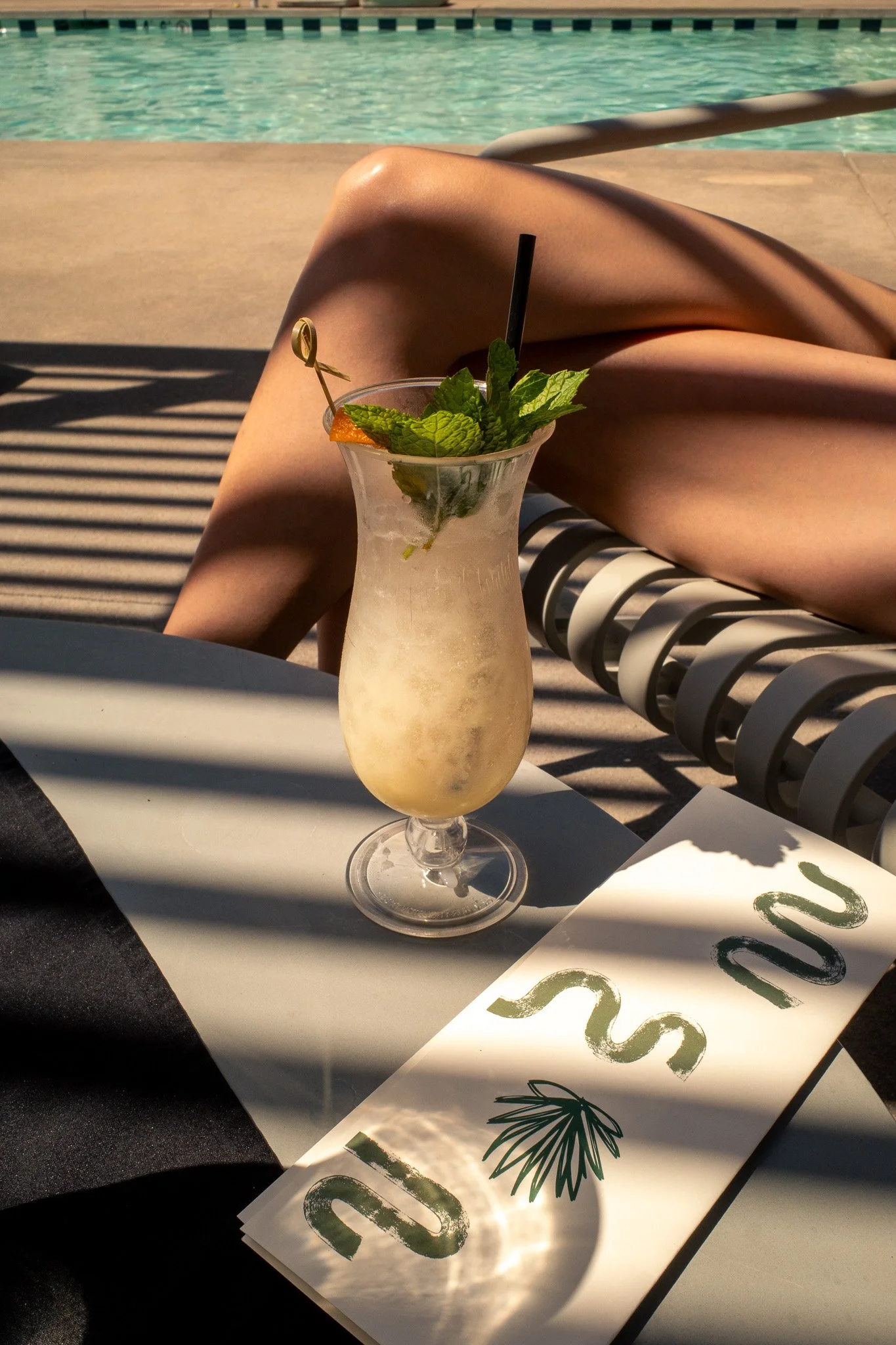 A tropical cocktail with mint leaves, an orange slice, and a straw, placed on a table near a swimming pool with a person lounging nearby.