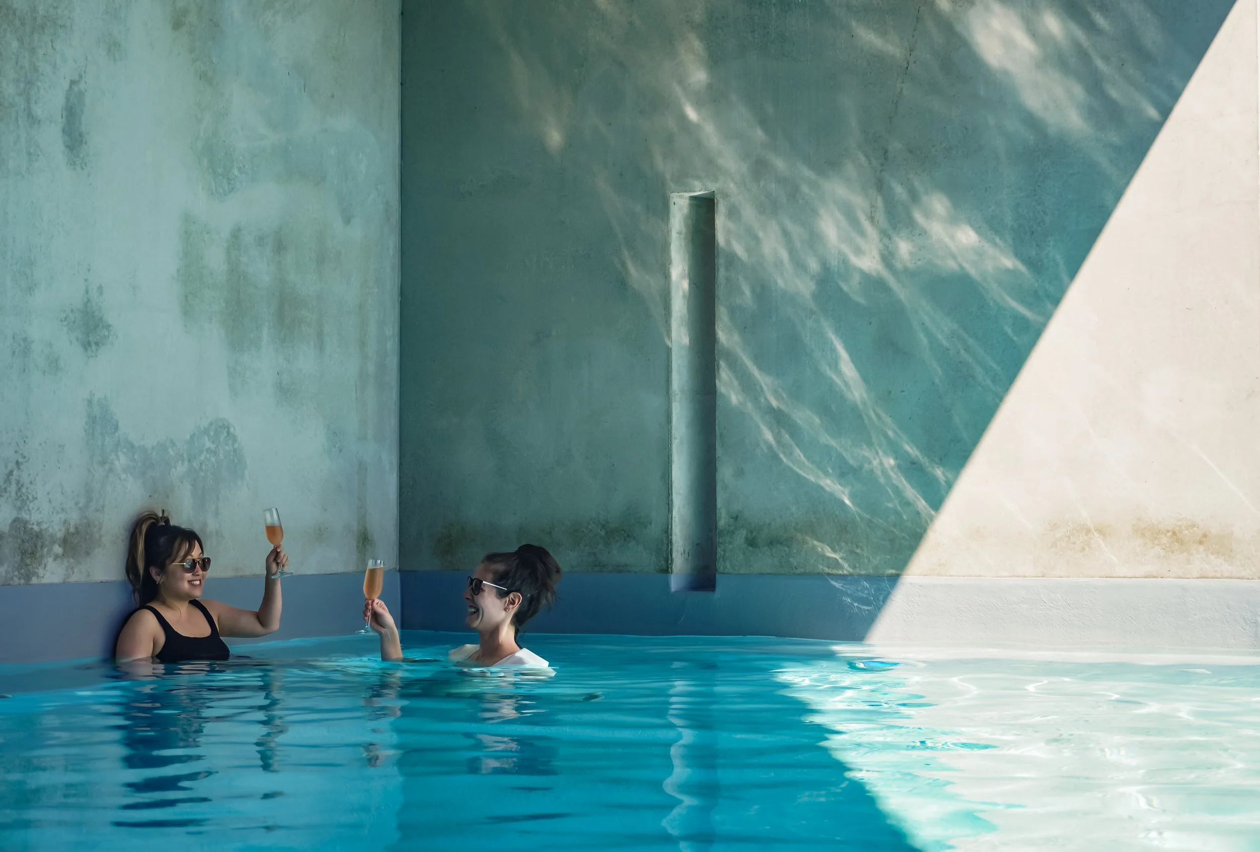 Two women wearing sunglasses in a pool, celebrating with glasses of rosé