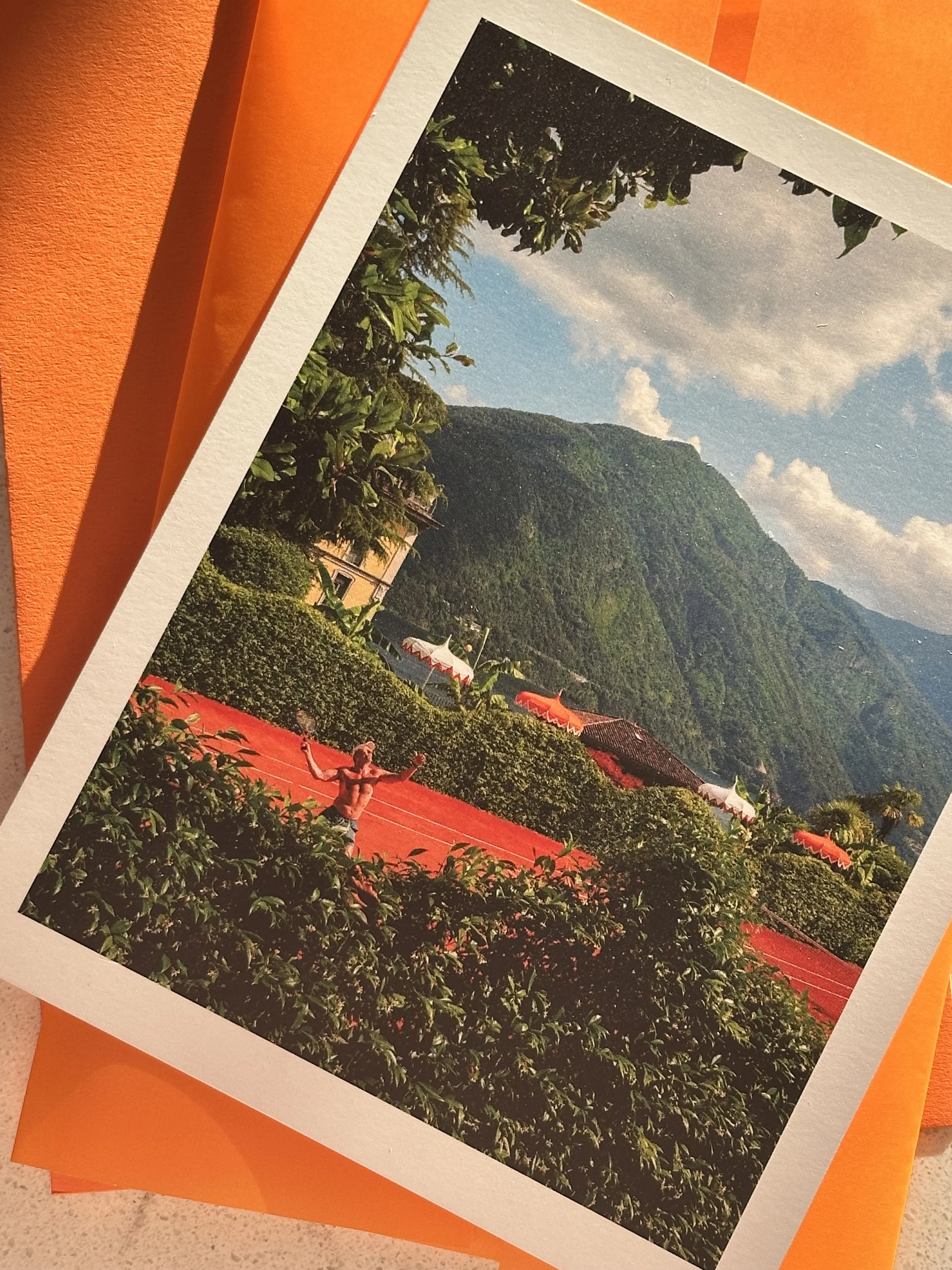 Photograph of a mountain landscape with clouds in the sky, greenery, and a person practicing yoga on a red outdoor court surrounded by bushes and decorative umbrellas.
