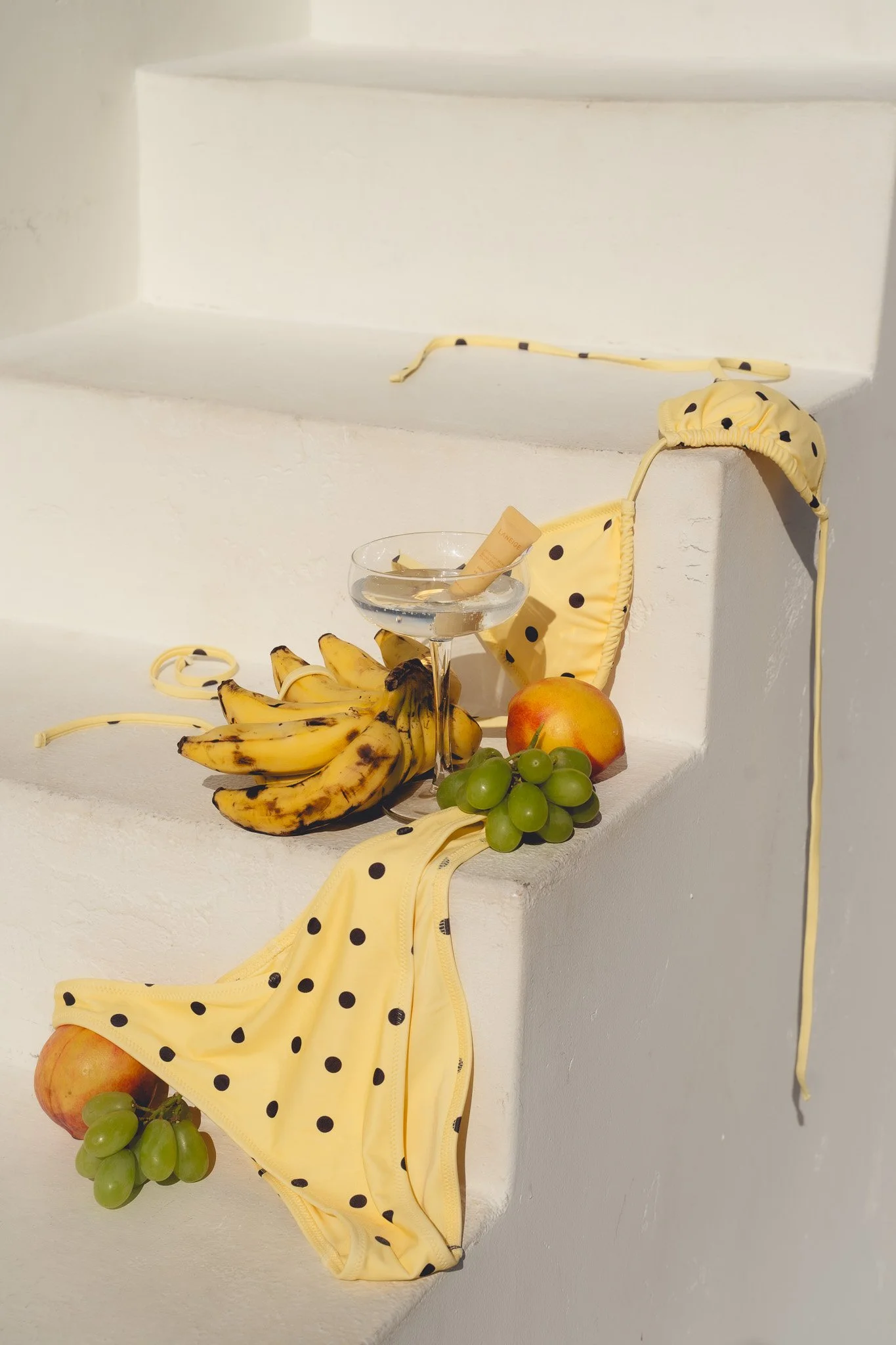 A still life arrangement on white stairs including yellow polka dot bikini, bananas, green grapes, apples, a drink in a martini glass, and a tube of lotion.