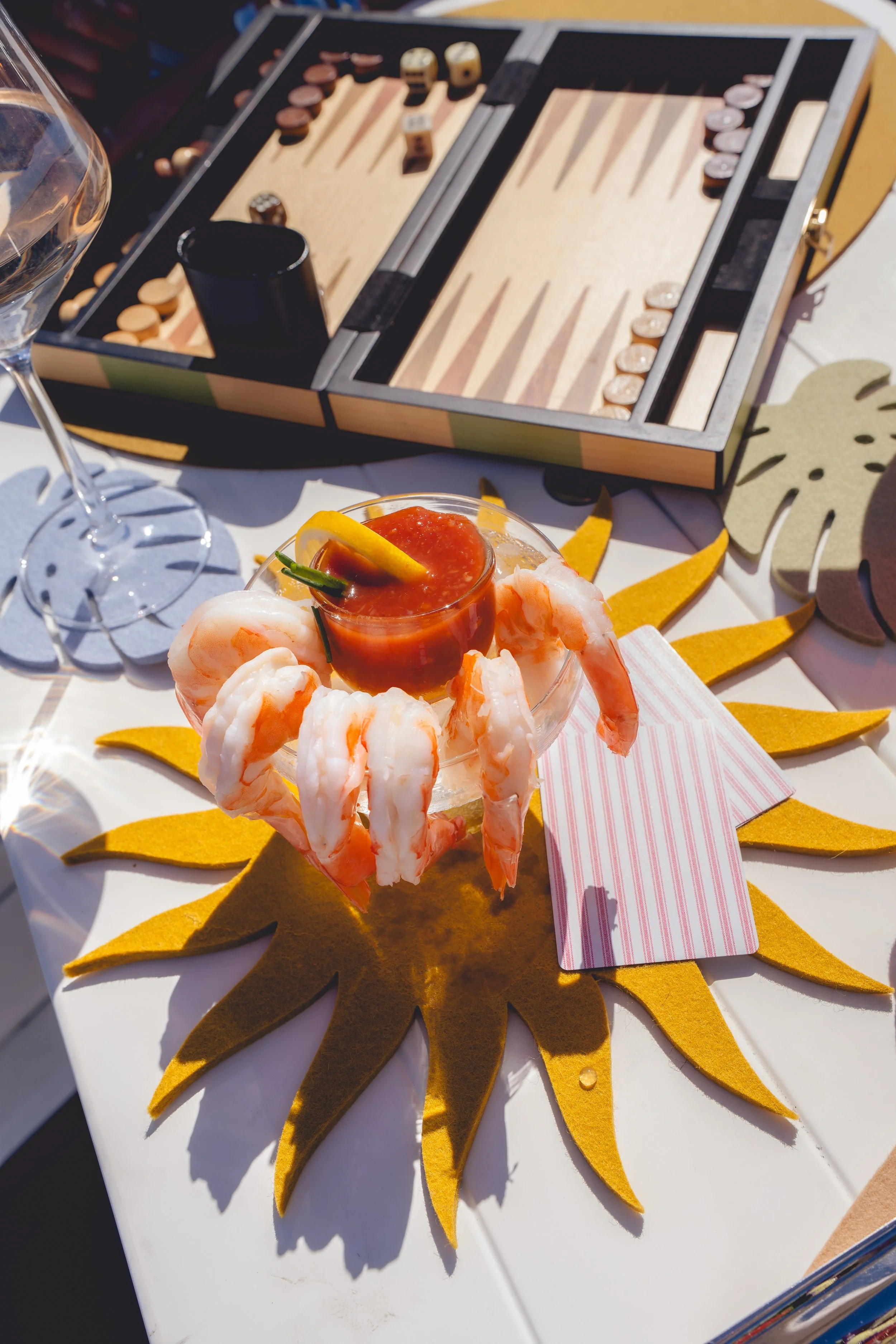 A table with a shrimp cocktail in a glass, surrounded by lemon wedges, on a sun-shaped felt placemat, with a backgammon game in the background.