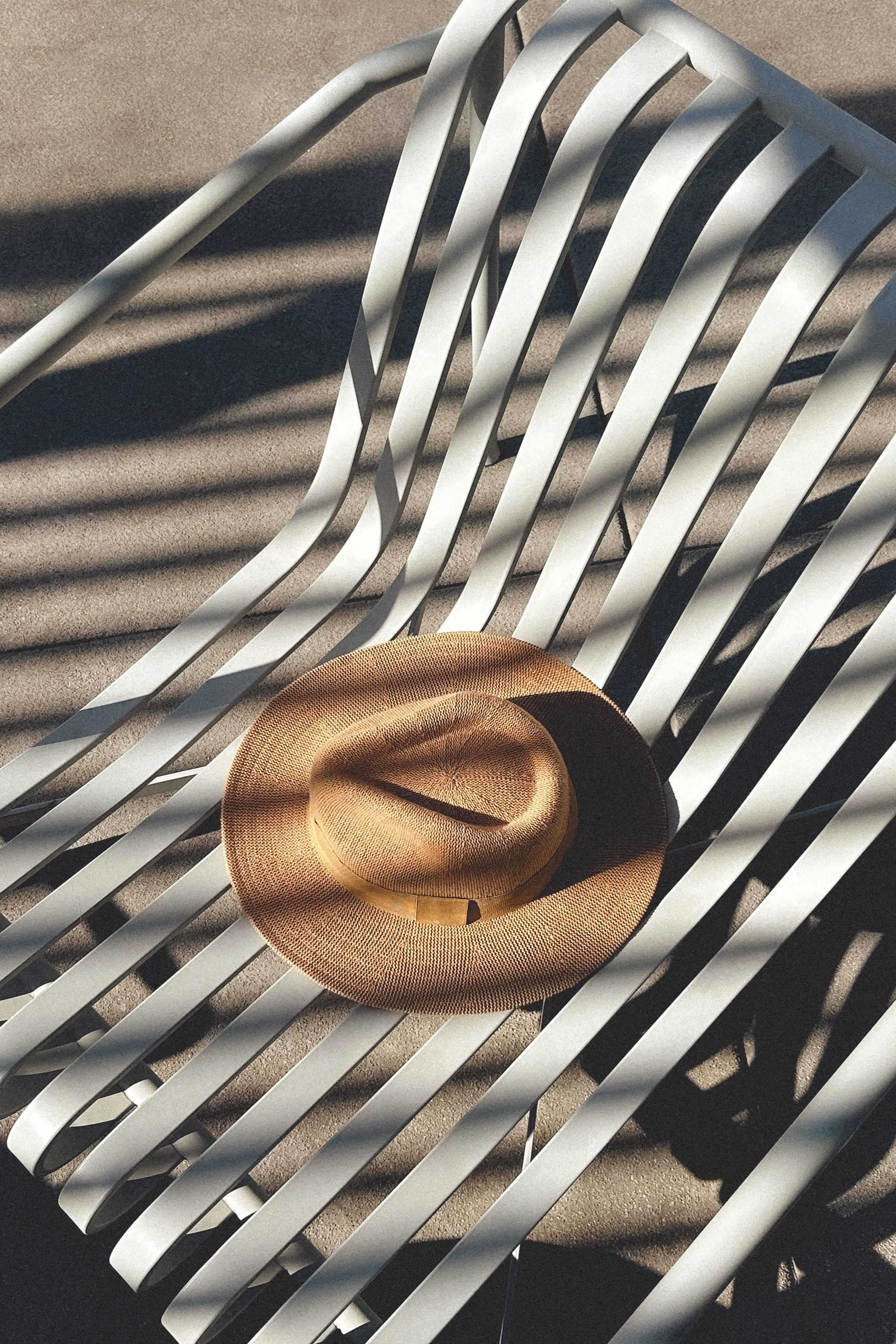 A straw sun hat resting on a white metal lounge chair on a sandy surface with shadows from nearby objects.
