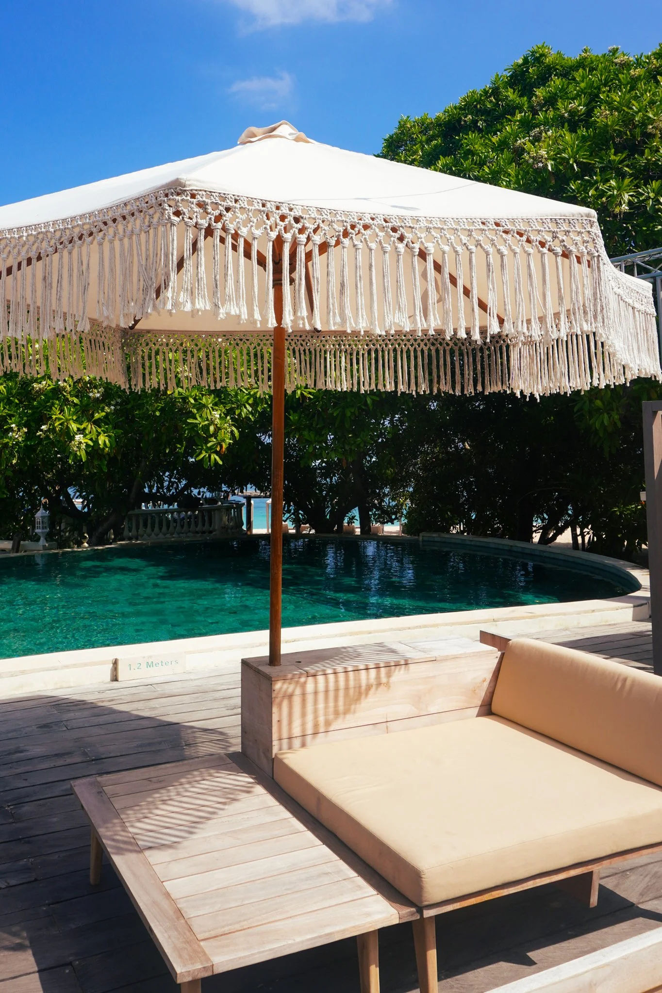 A sunlit poolside scene with a beige cushioned lounge chair, a large white fringed umbrella, and lush green trees in the background, overlooking the swimming pool.