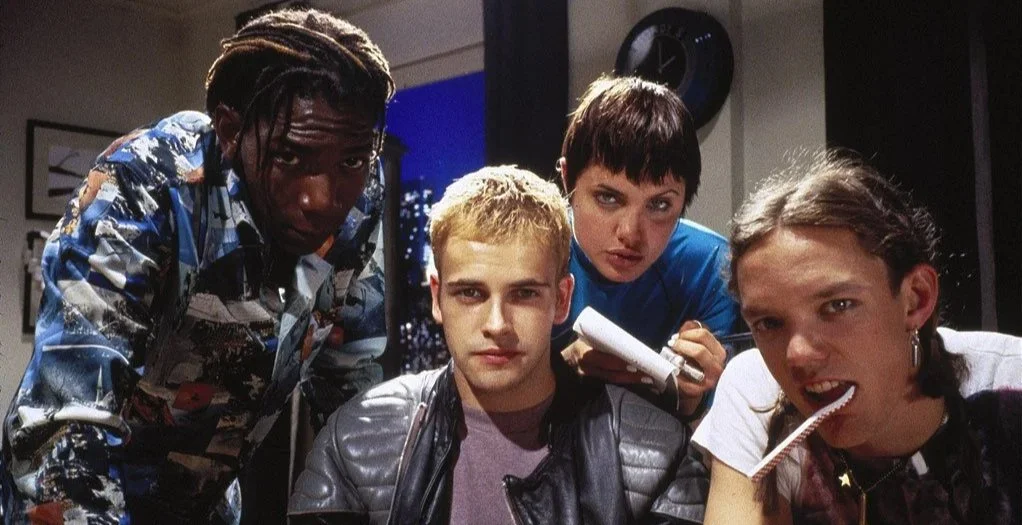 Group of five young people gathered around, looking at the camera with serious expressions, inside a room with wall art and a clock, one person biting a lollipop.