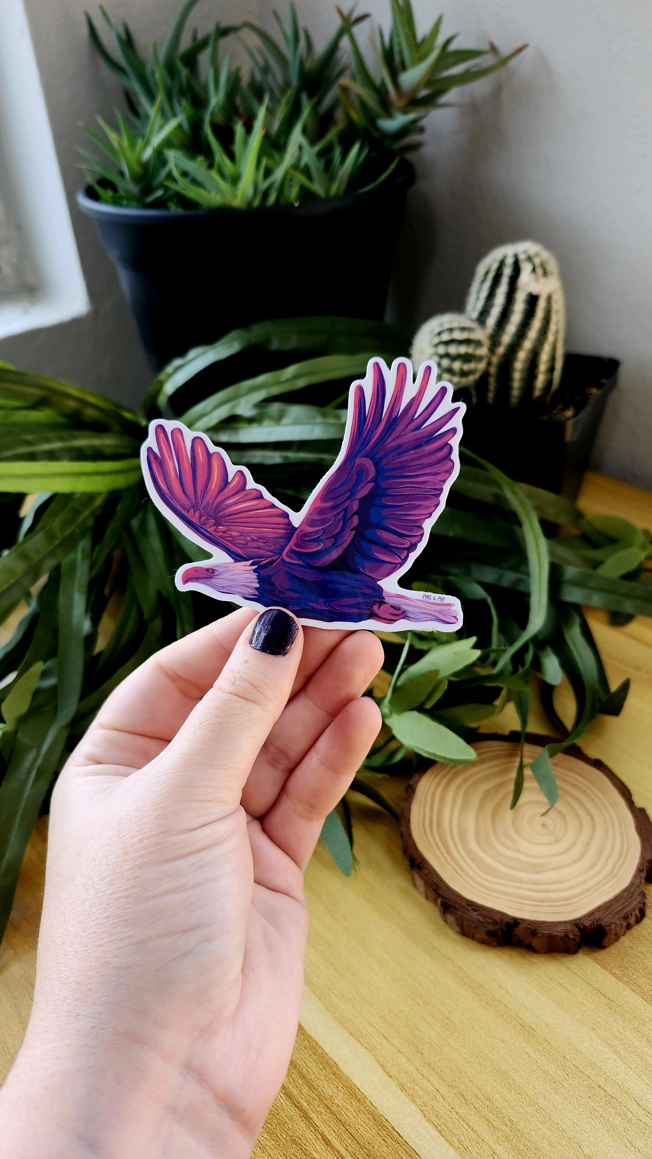 A hand holding a sticker of a bald eagle in flight with bold purple and pink colors