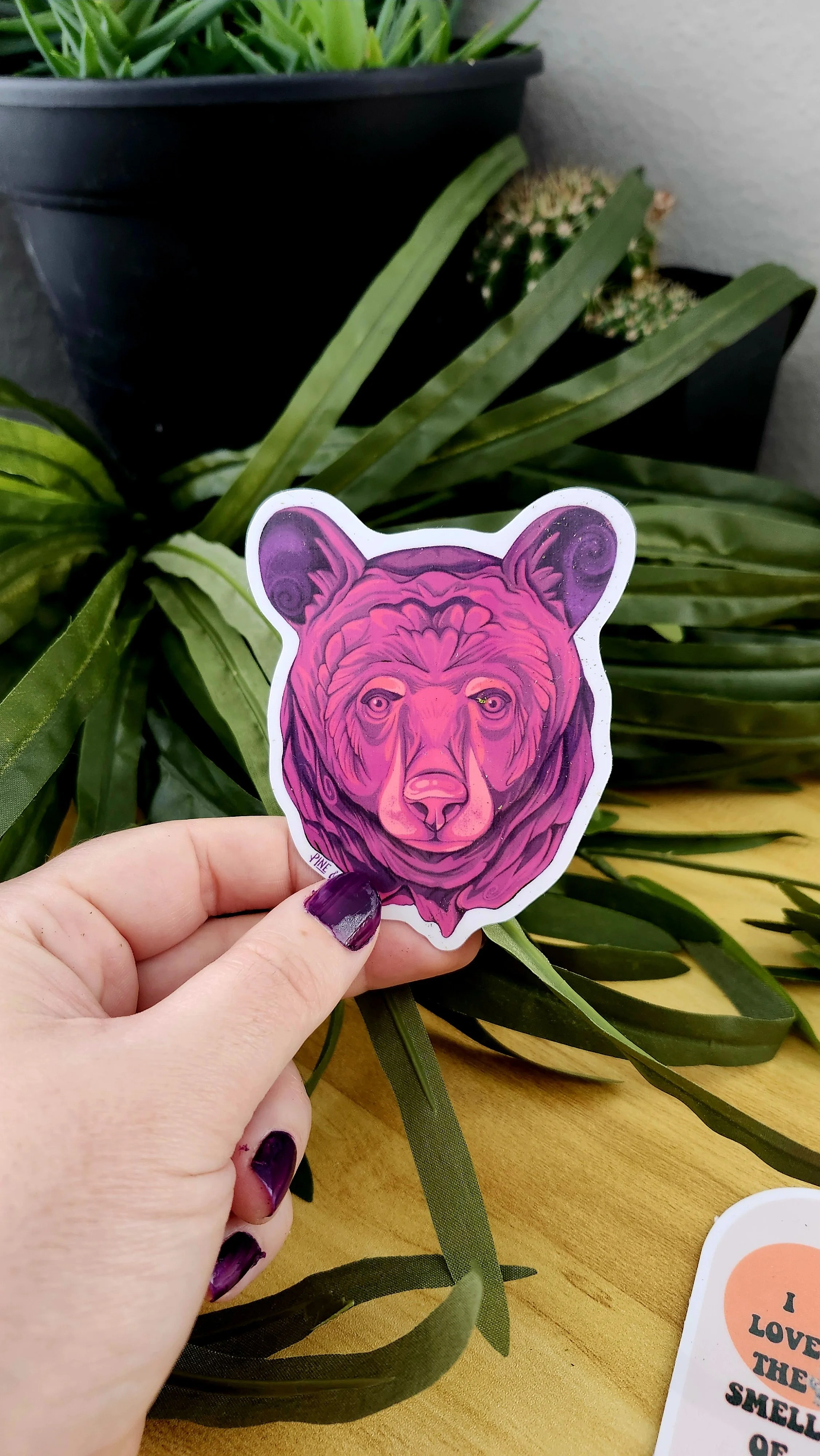 a hand holding a bear face sticker made up of purple, mauve, pink, and peach colors