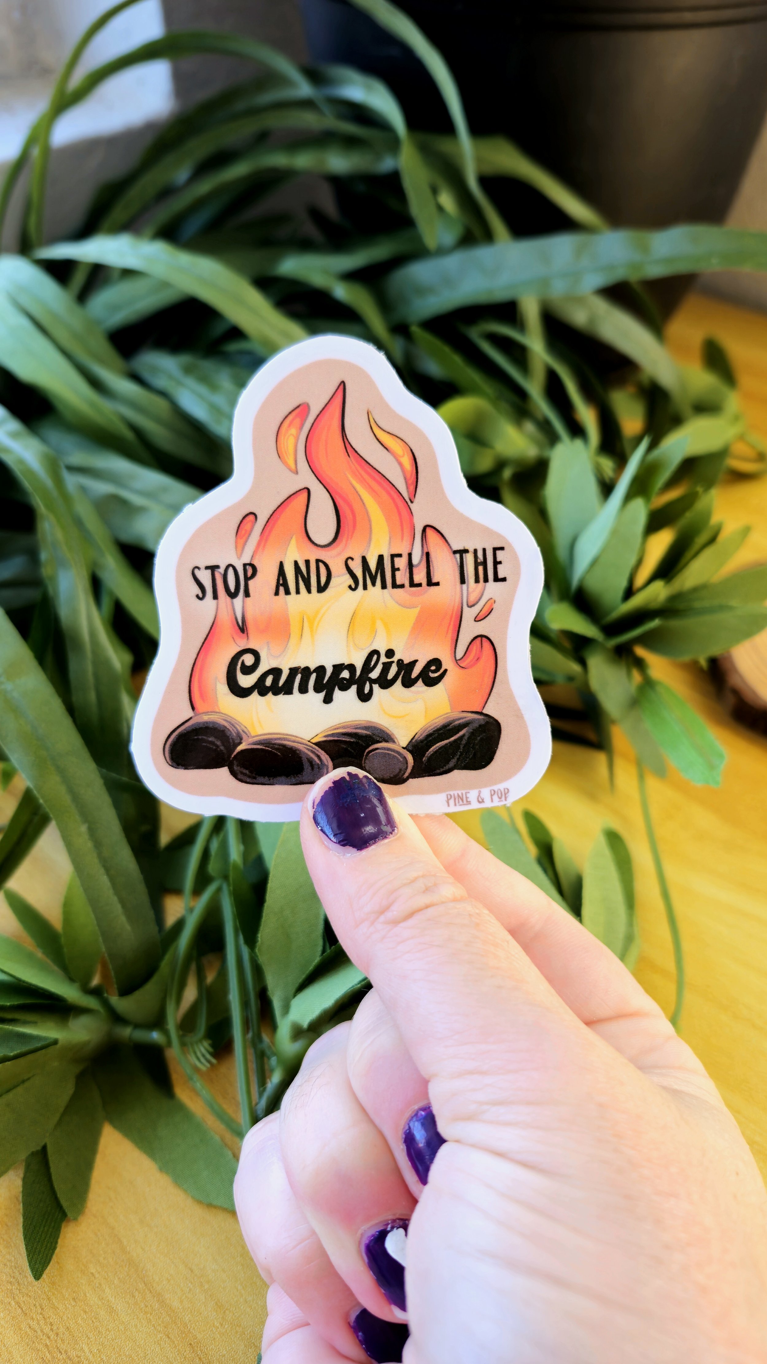 A hand holding a sticker of a flaming campfire with the words “Stop and Smell the Campfire” written in the middle.