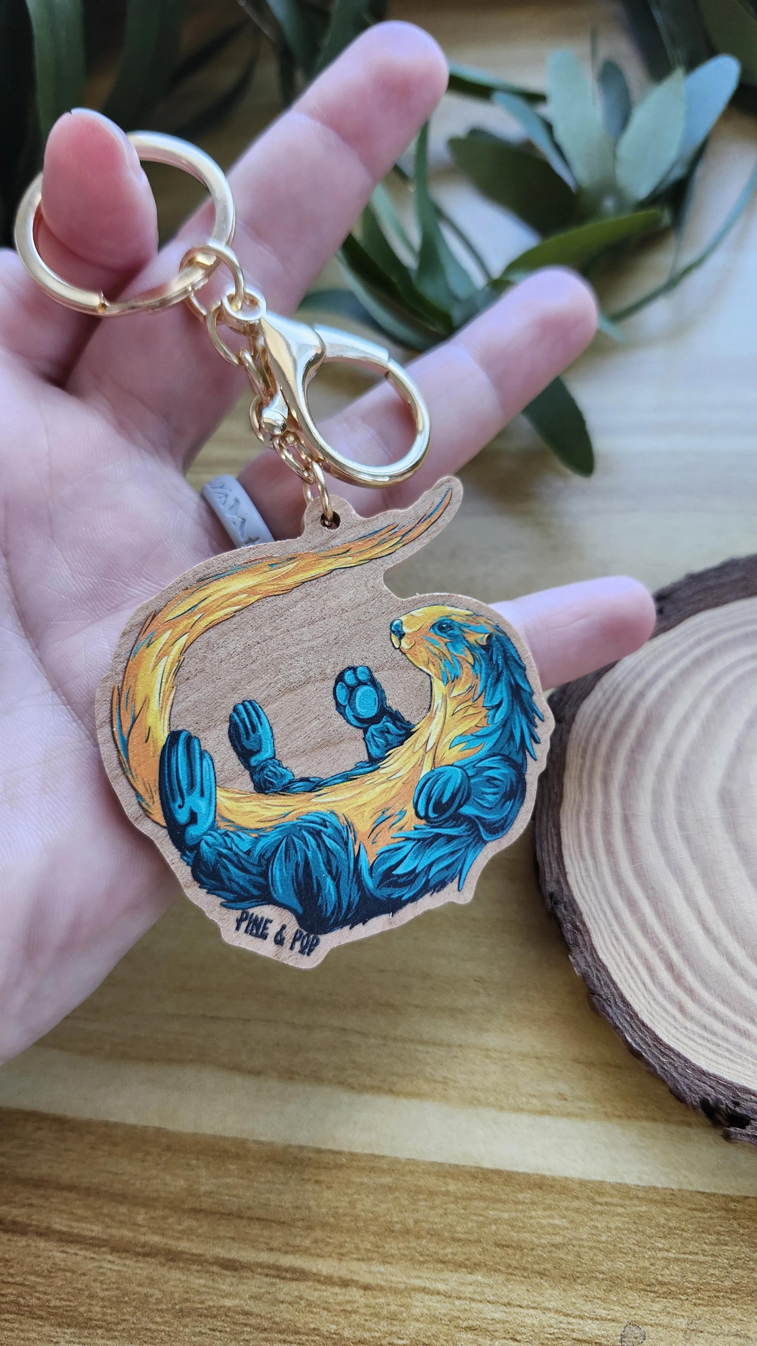hand holding a wooden keychain of an otter with orange, yellow, and teal colors with gold hardware