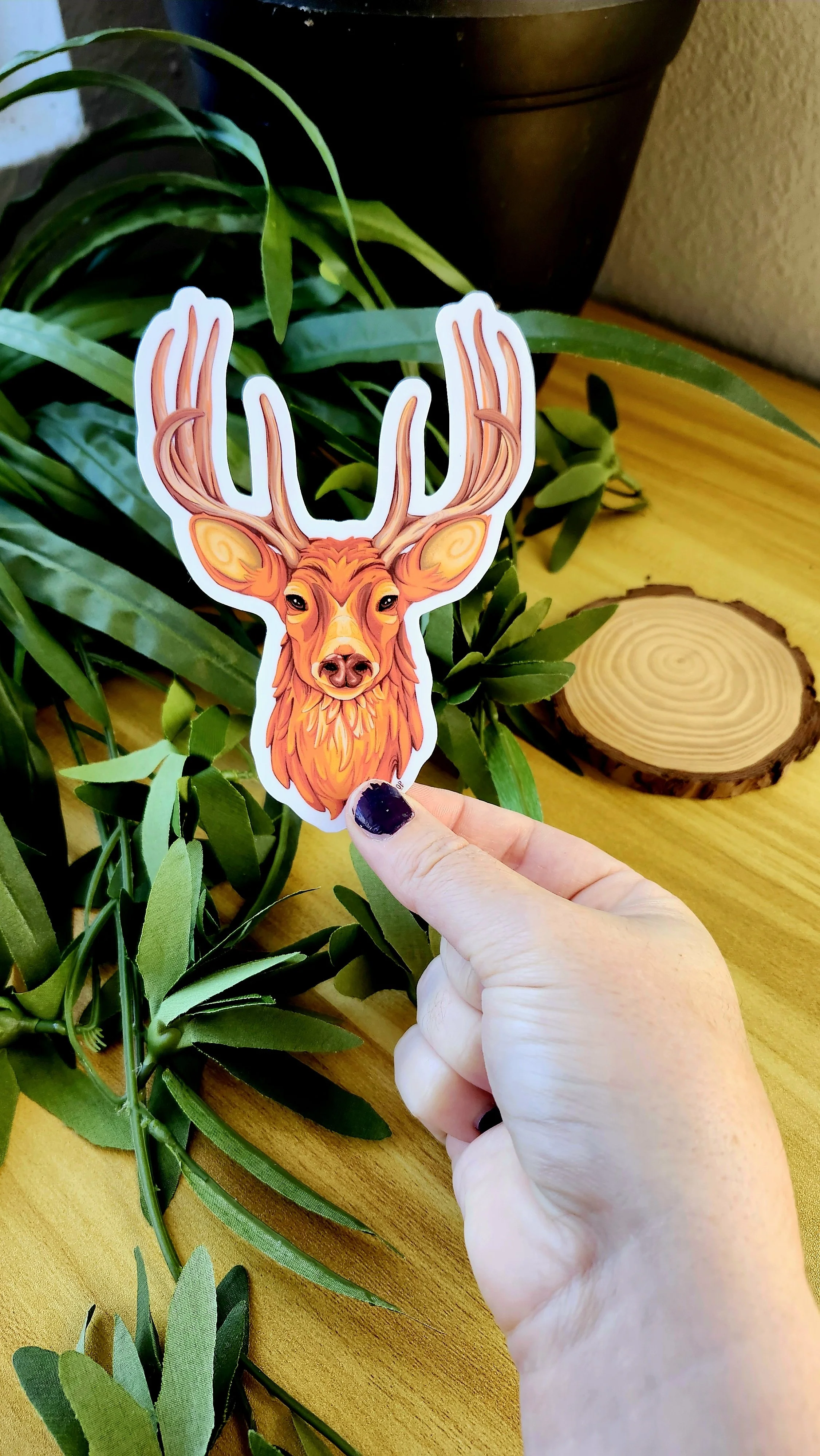 a hand holding a sticker of a deer head with antlers using yellow, oranges, and browns in the design.