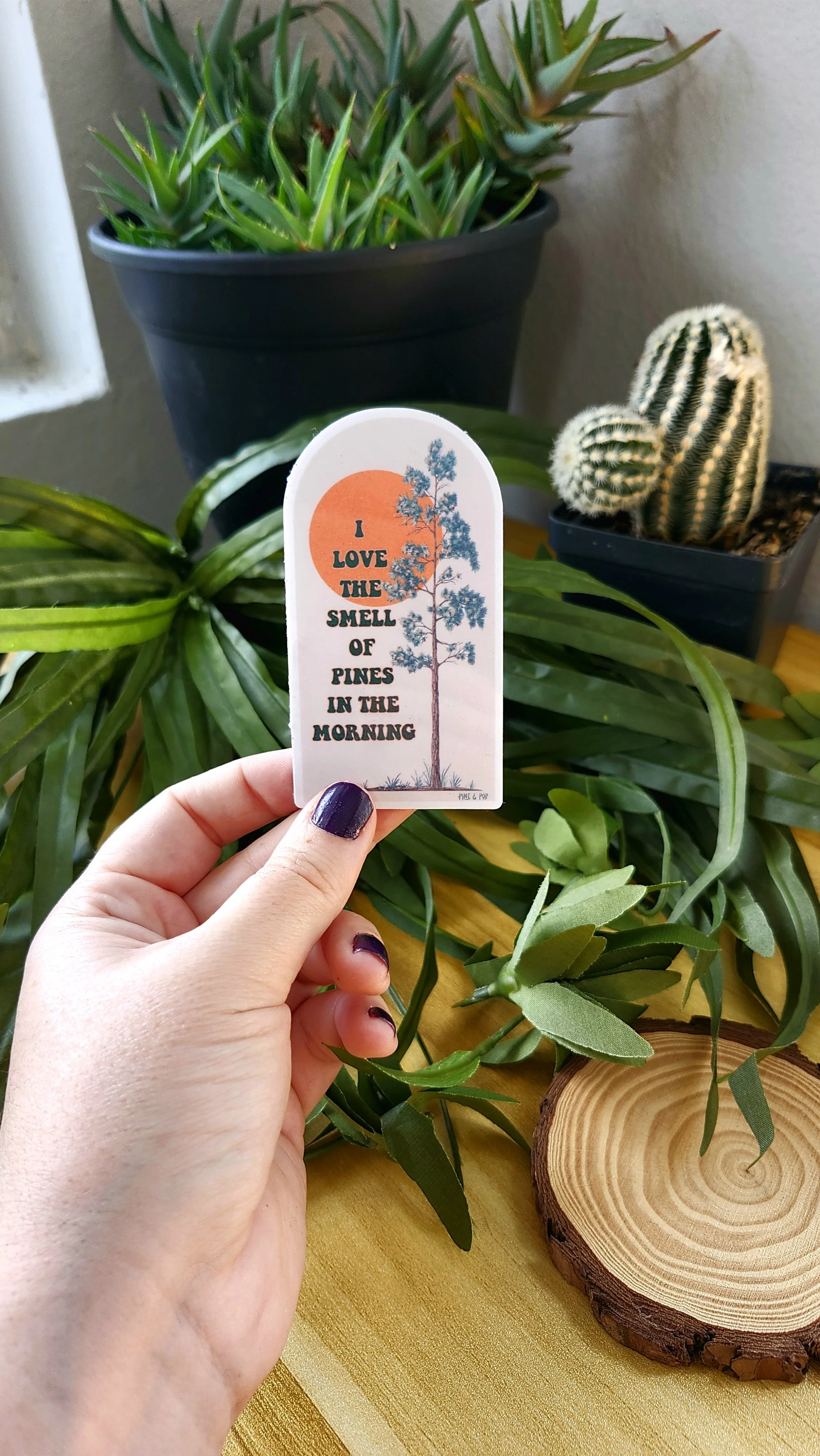 a hand holding a sticker with a pine tree on the right and an orange sun with a light brown/cream background  and I love the smell of pine in the morning written on it