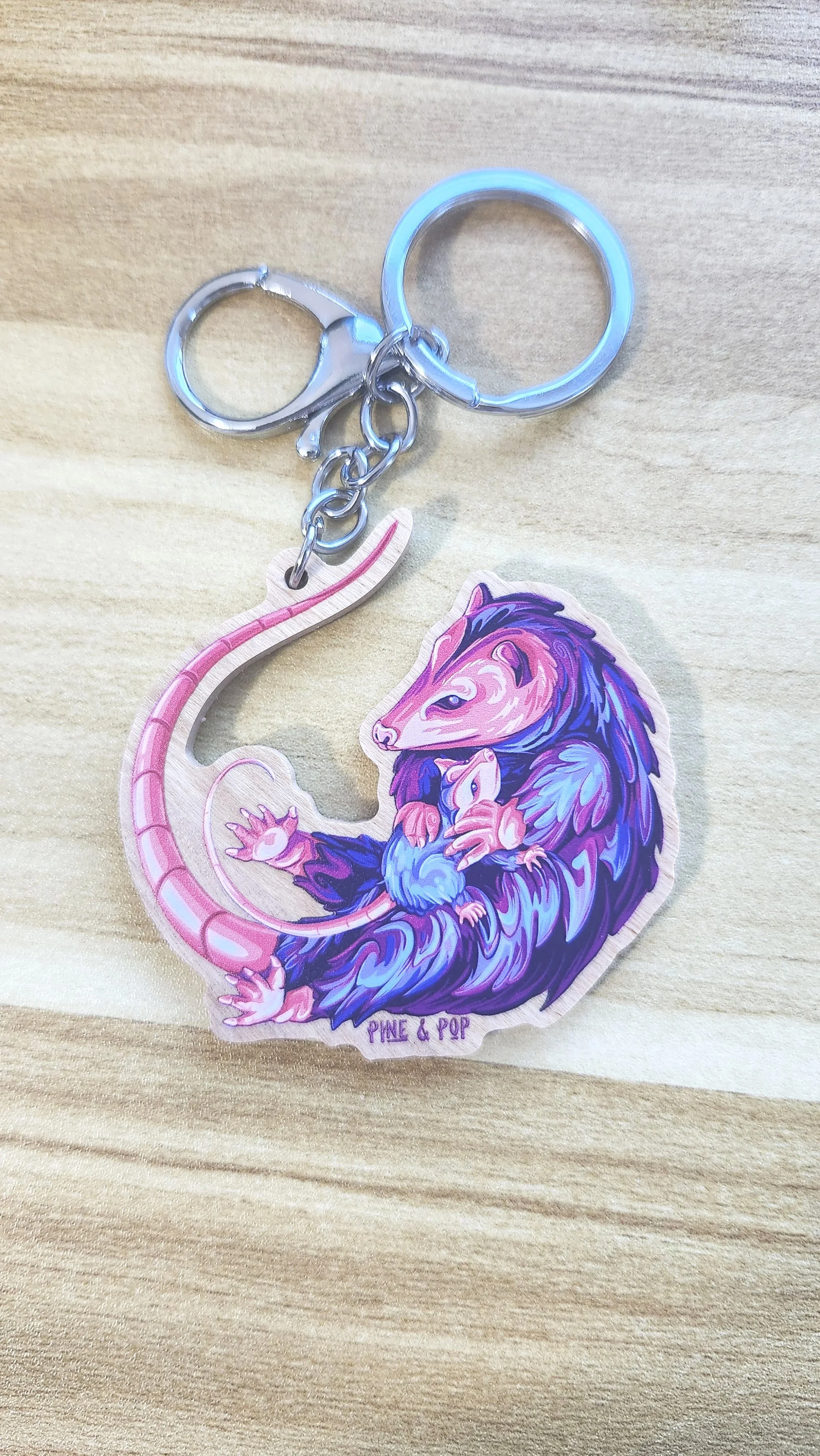 wooden keychain of opossum designed with bright blue, purple, pink, and coral colors