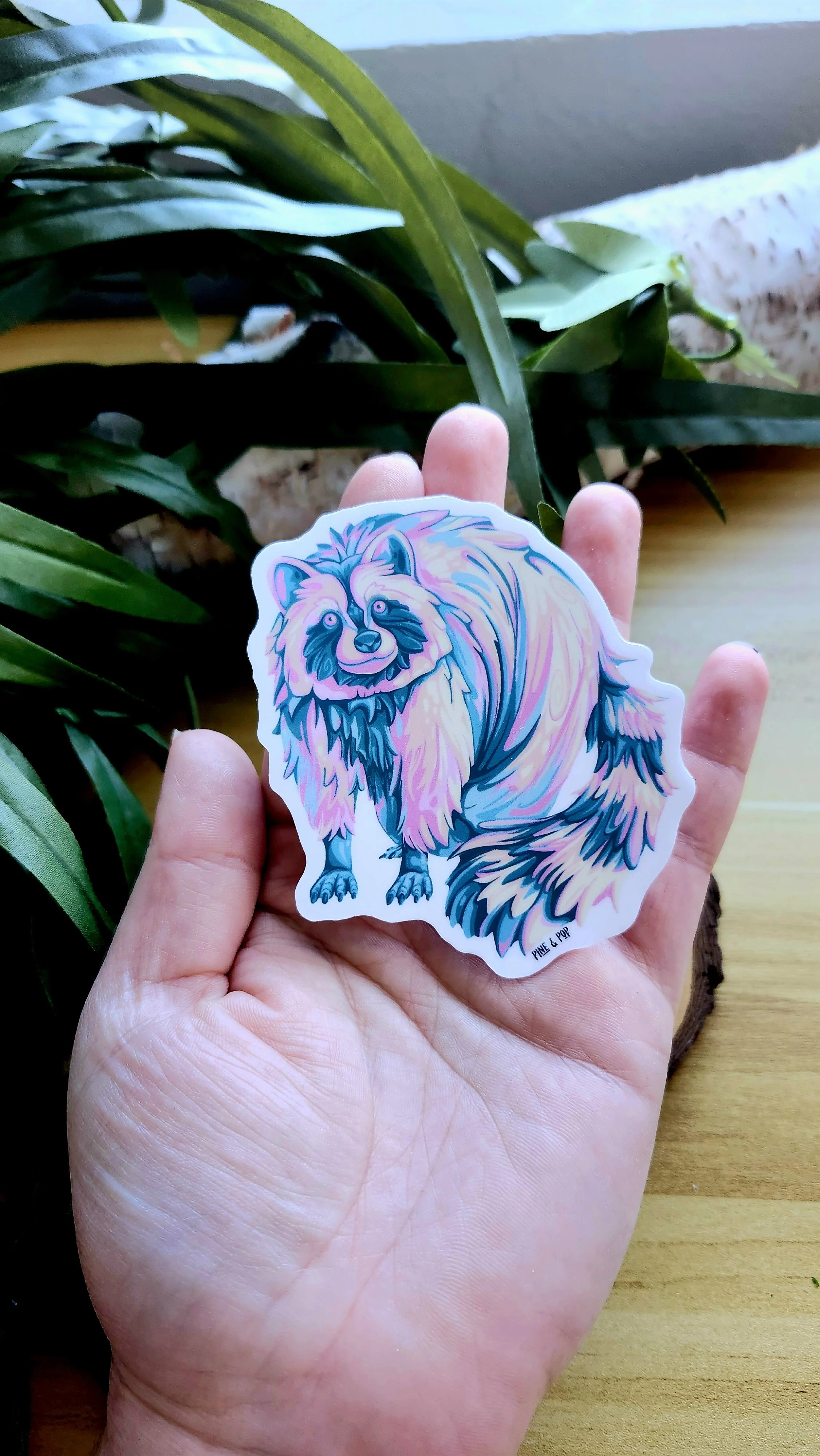 a hand holding a pastel colored raccoon sticker