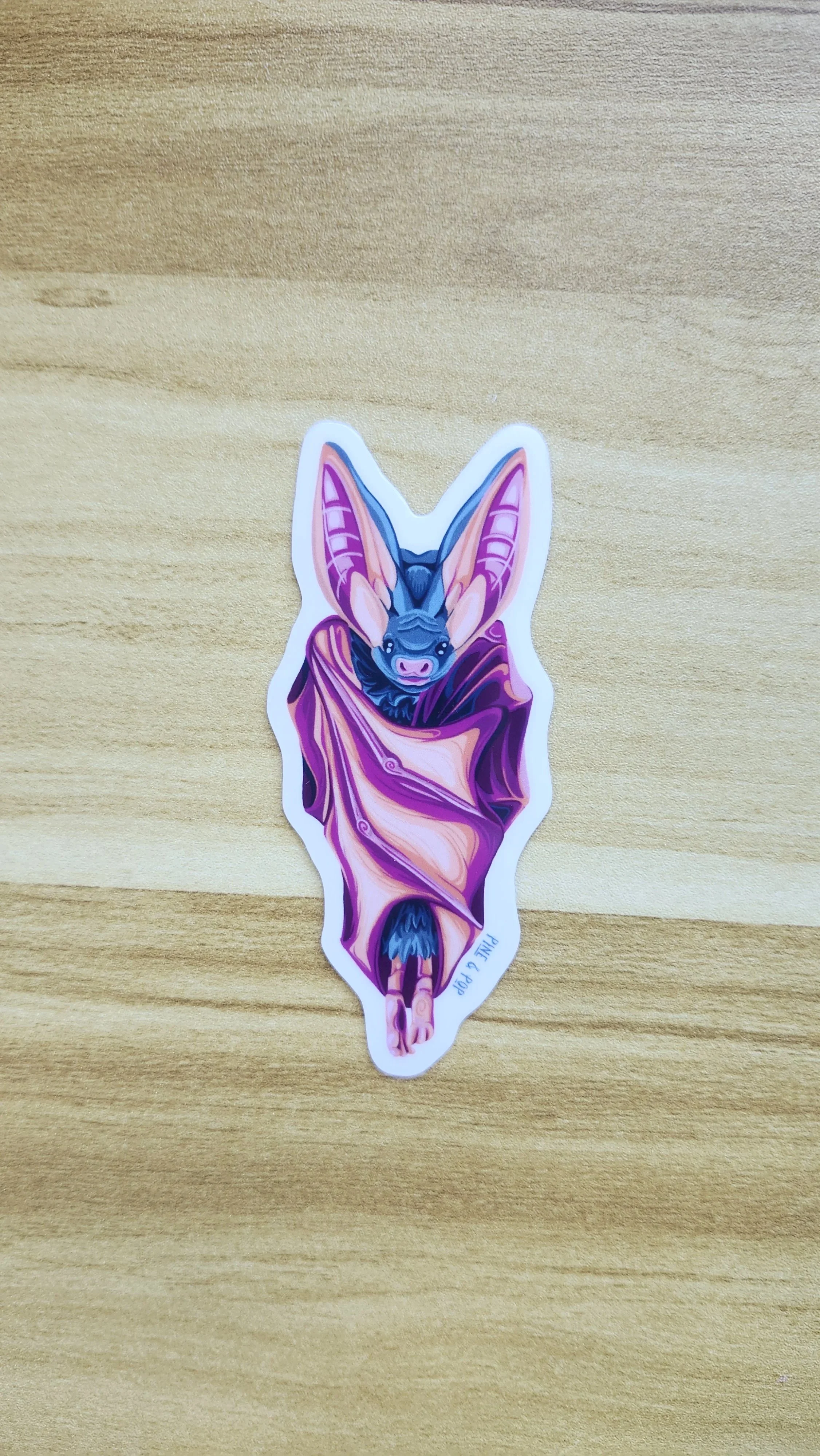 sticker of bat hanging with its wings wrapped around its body designed with orange, teal, pink, and purple colors