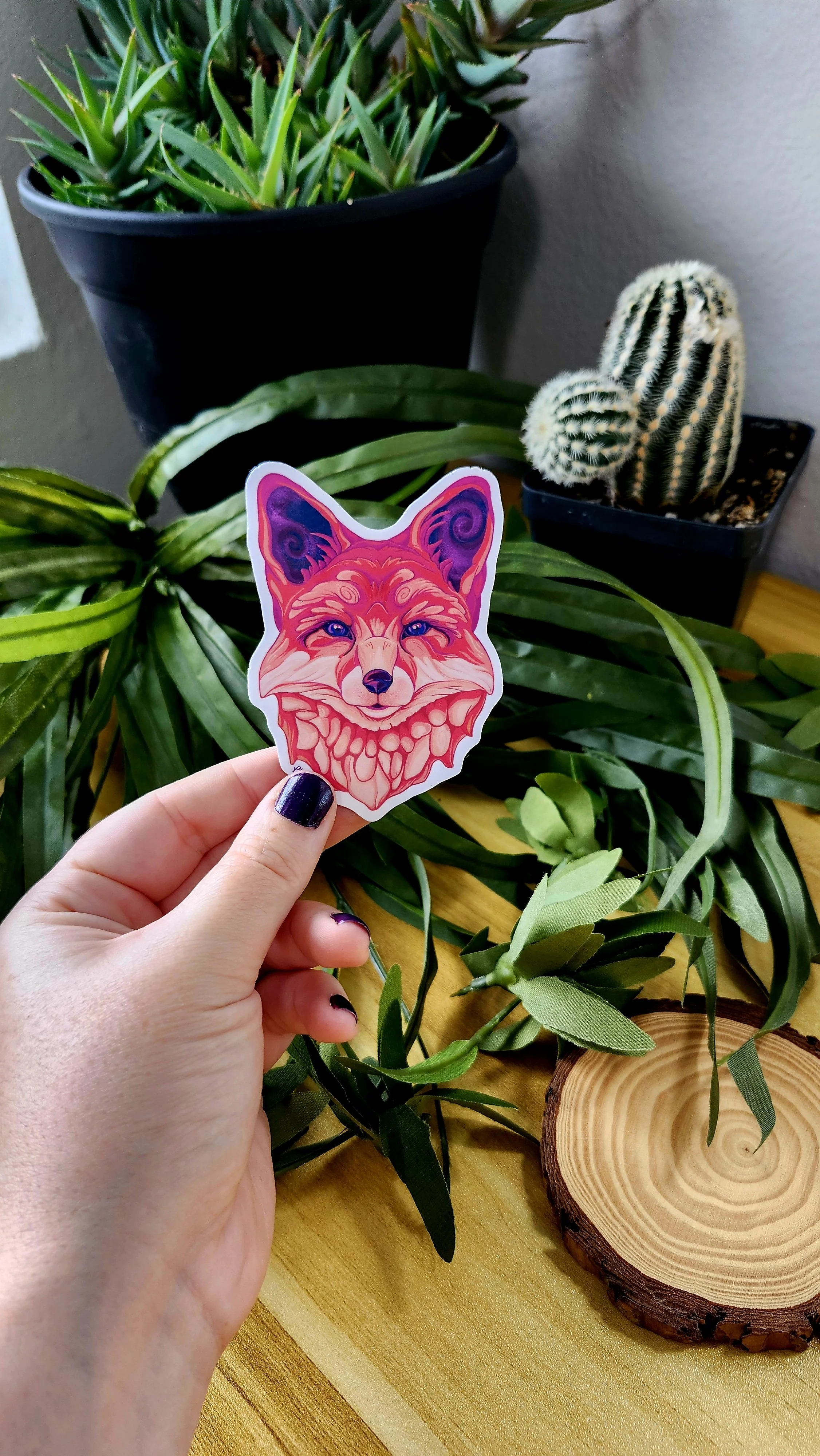 a hand holding a sticker of a fox head with red, orange, cream, and purple colors used to create the design