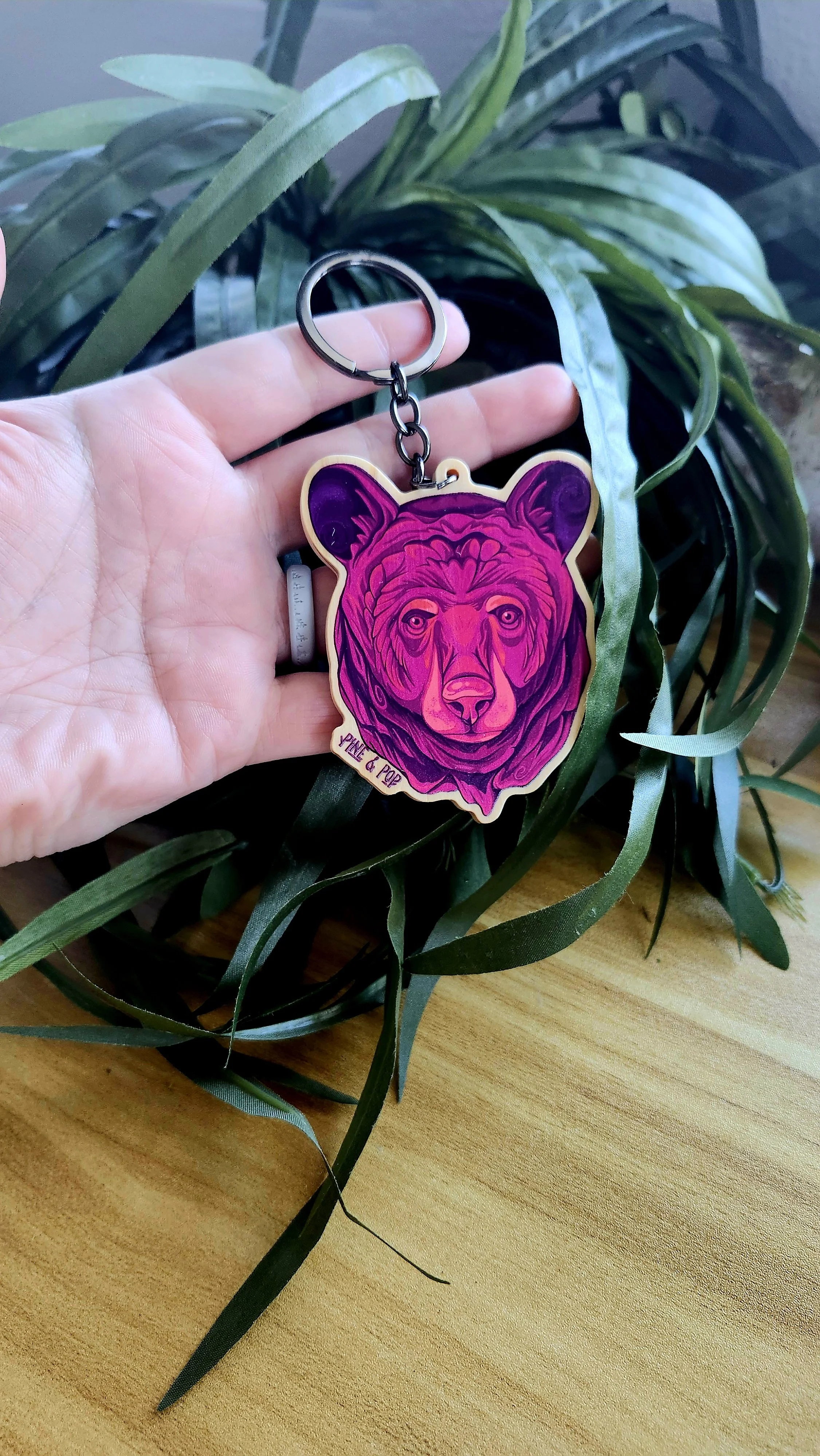 keychain of black bear face with purple, mauve, and peach tones with a gunmetal grey chain and keyring in someone's hand