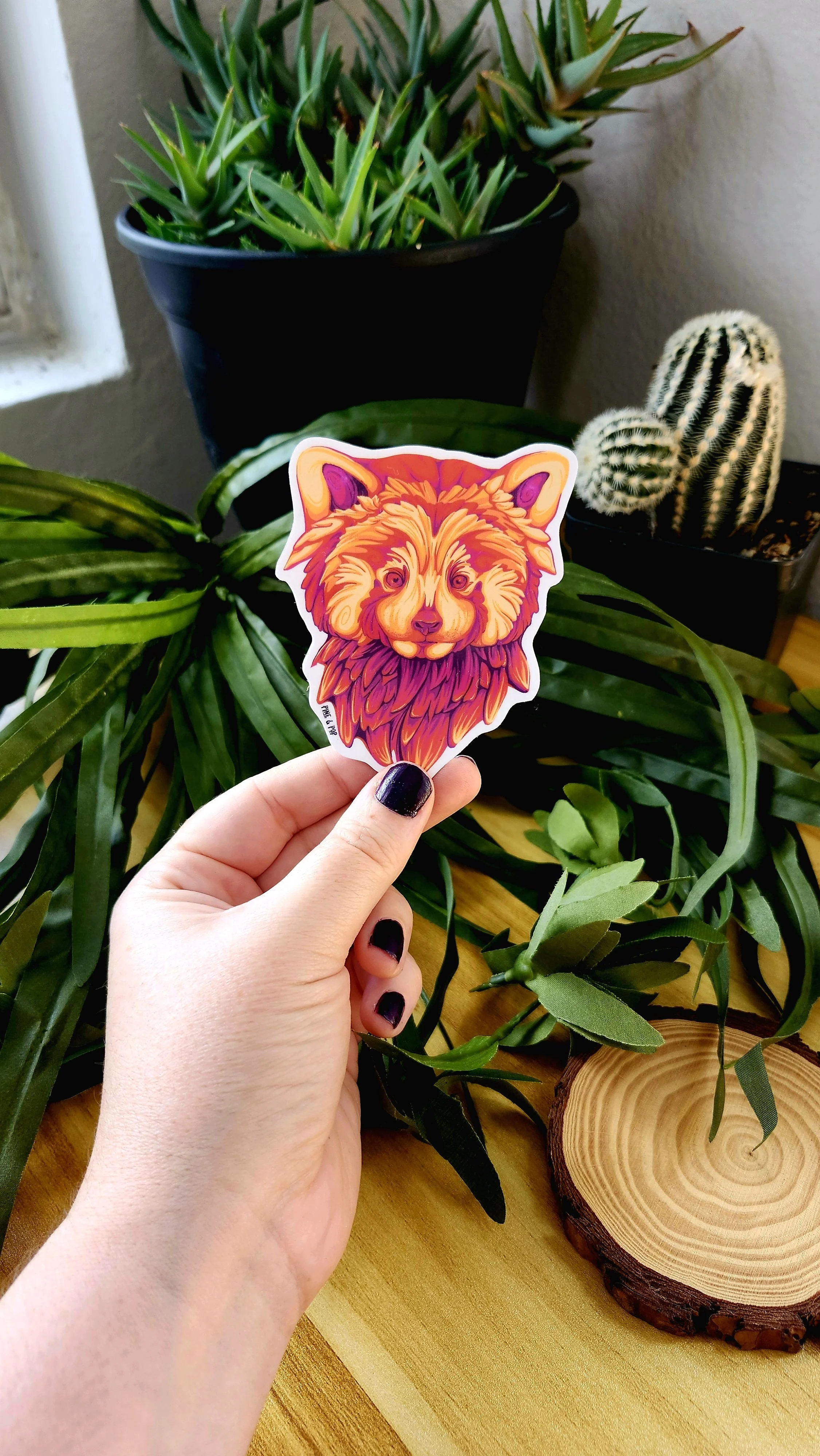 a hand holding the sticker of a mulit-colored red panda face