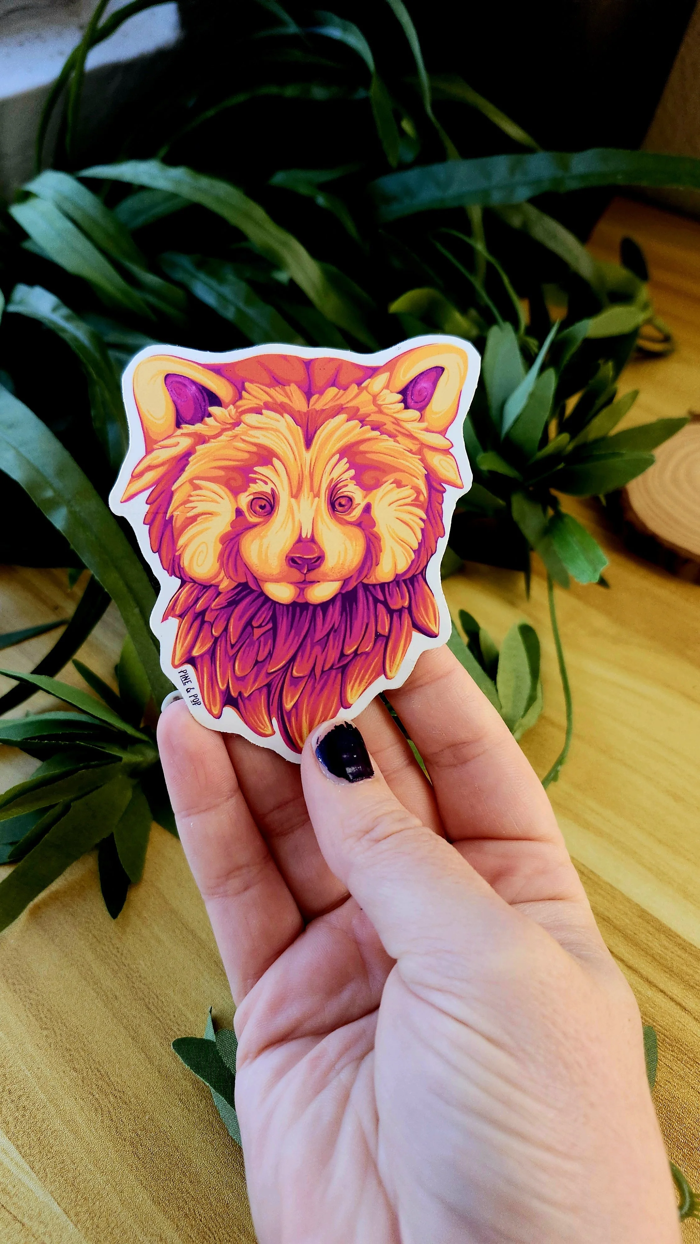 a hand holding a red panda face magnet with reds, oranges, yellows, and fuschia colors colors