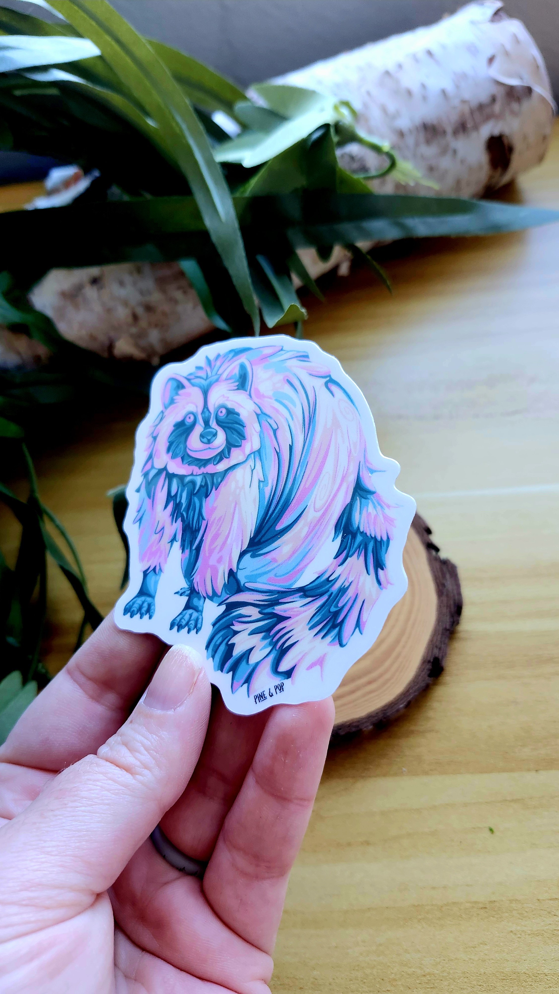 another view of a hand holding a pastel colored raccoon sticker