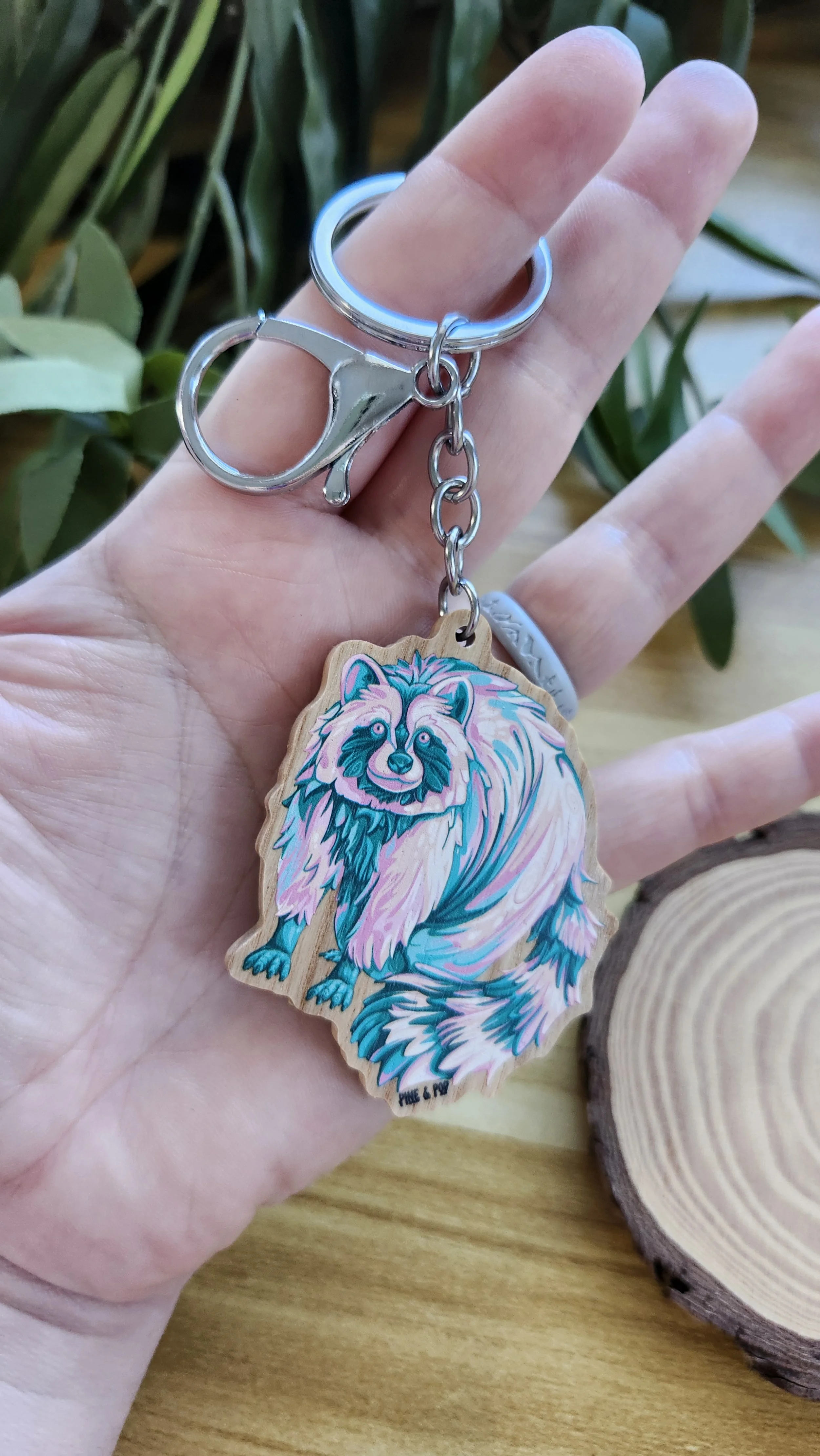 hand holding a wooden keychain of a raccoon with colorful pastel colors of light orange, light yellow, light pink, and teal