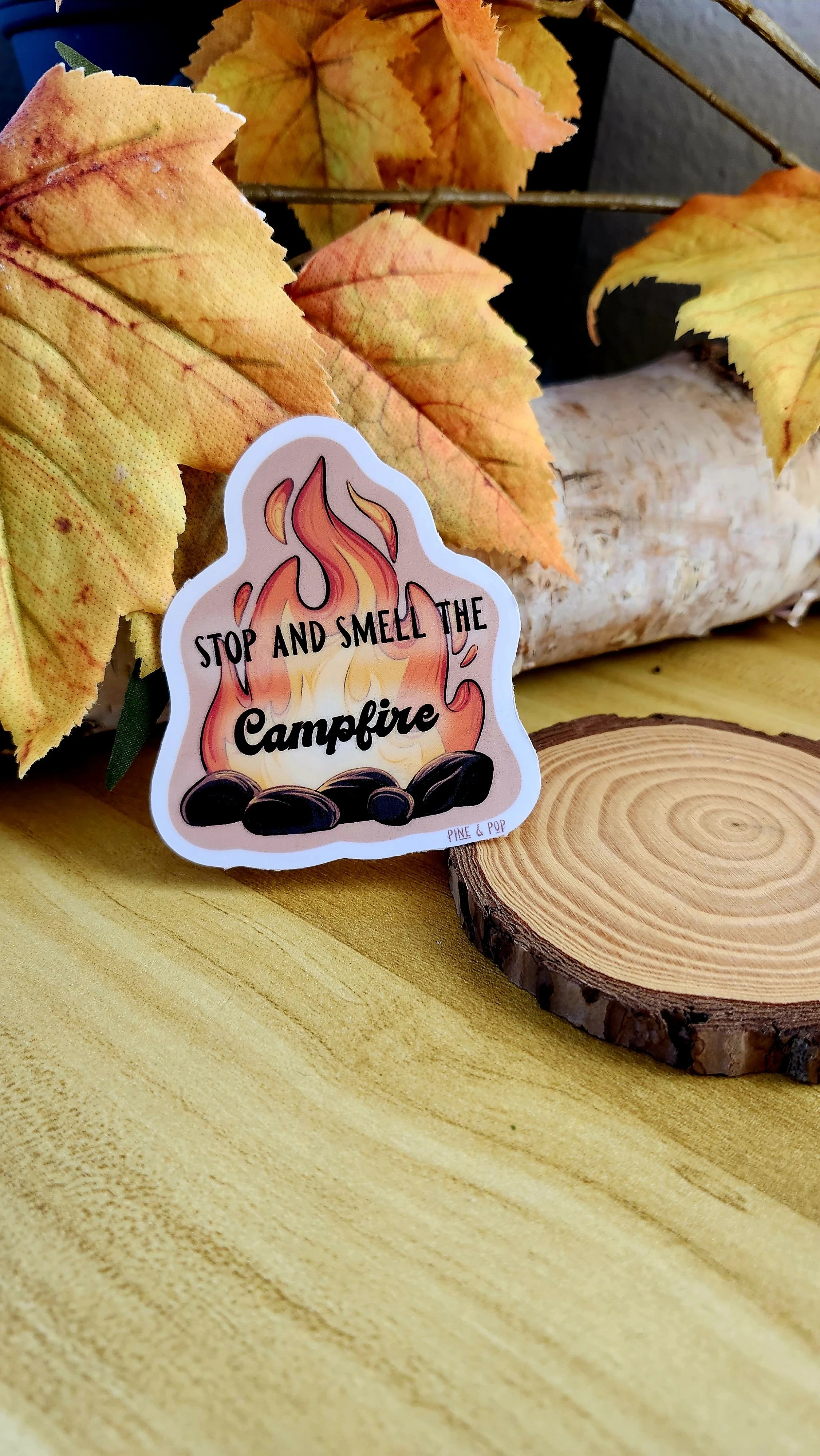 a sticker of a flaming campfire with the words “Stop and Smell the Campfire” written in the middle, laying against a tree section