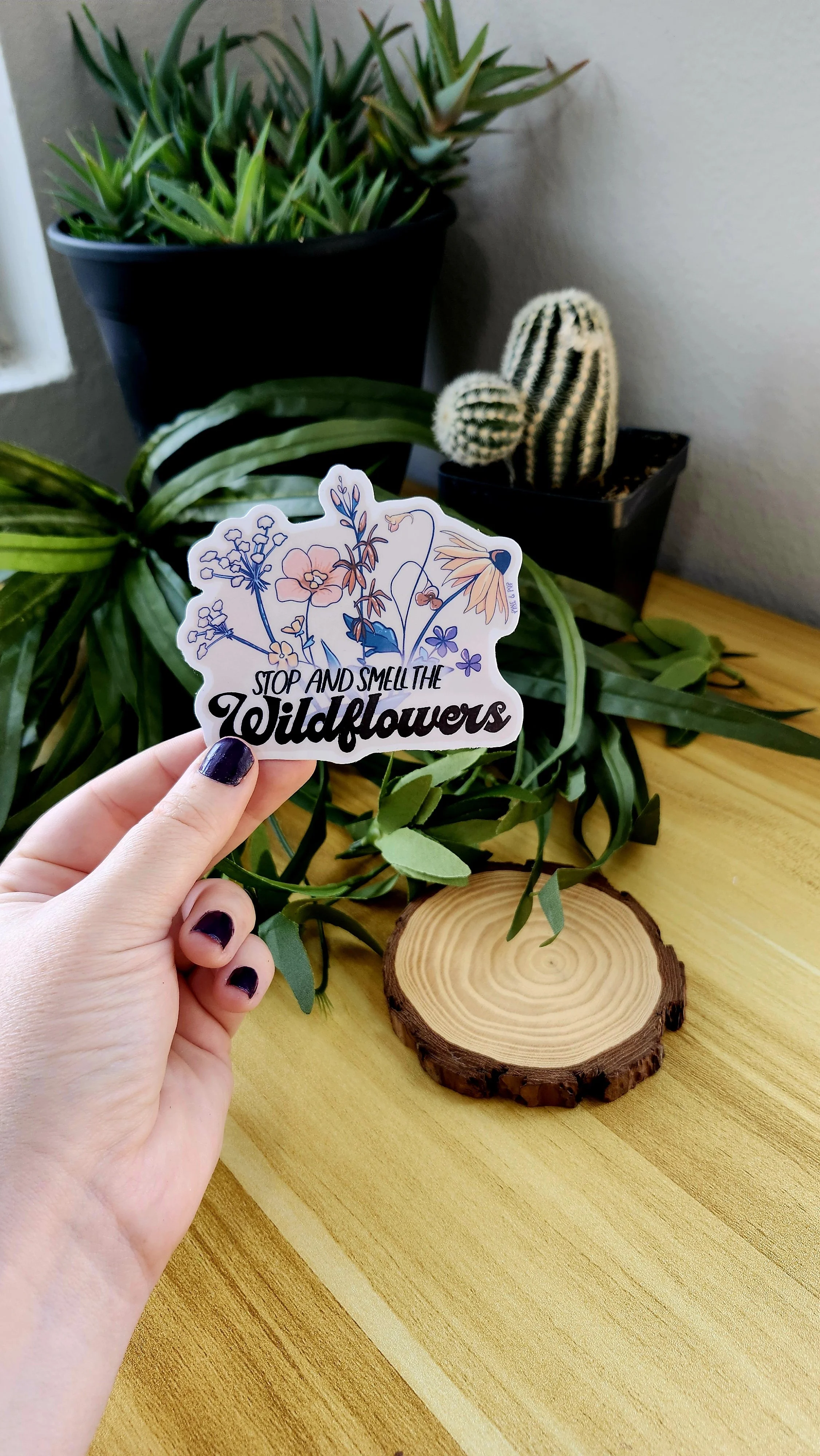 a hand holding a sticker depicting various wildflower species and the words Stop and Smell the wildflowers