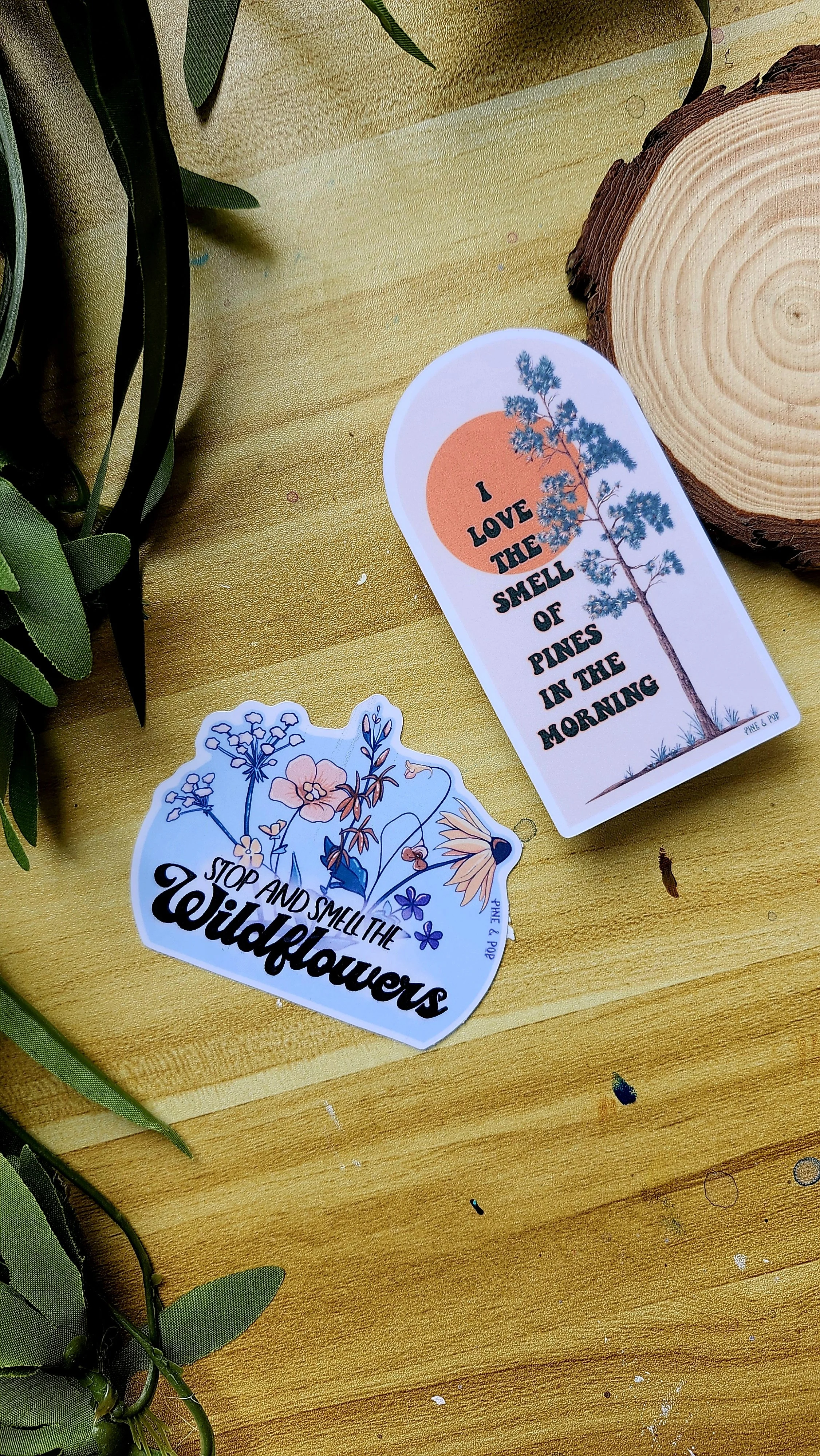 a I love the smell of pine in the morning sticker and a stop and smell the wildflowers sticker laying on a wooden table
