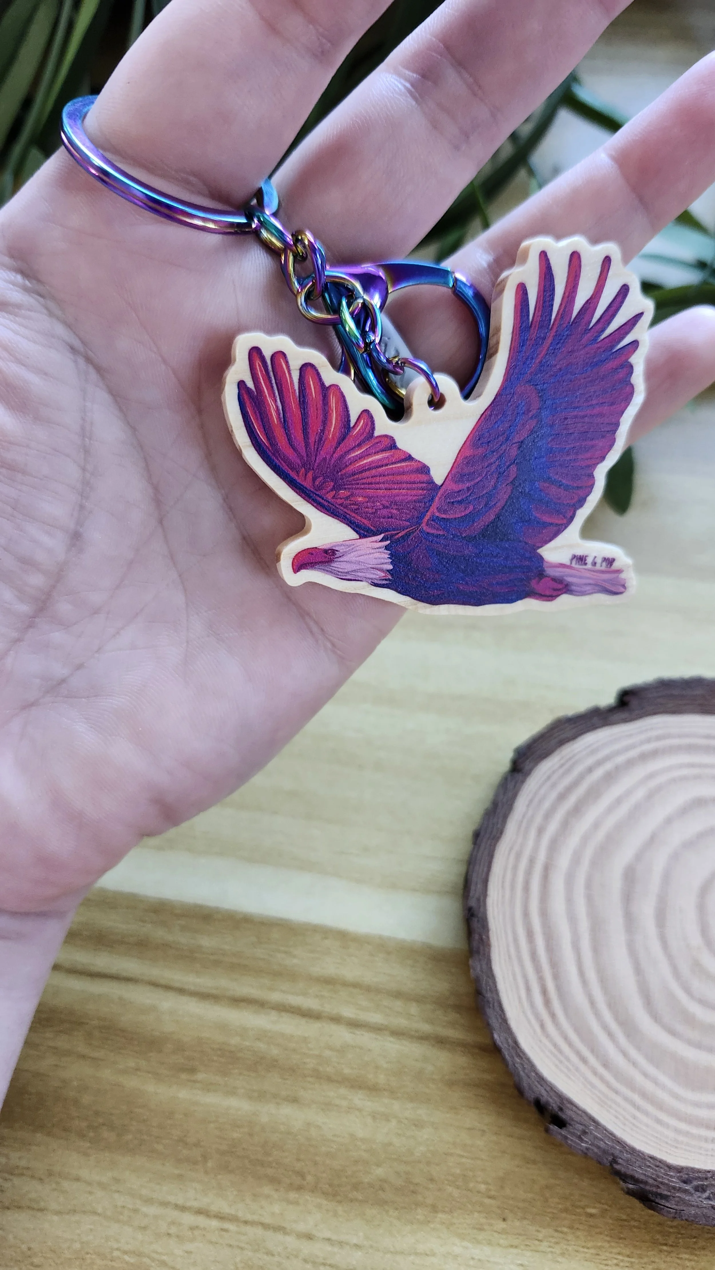 a hand holding a wooden keychain of a bald eagle in flight with bold purple and pink colors