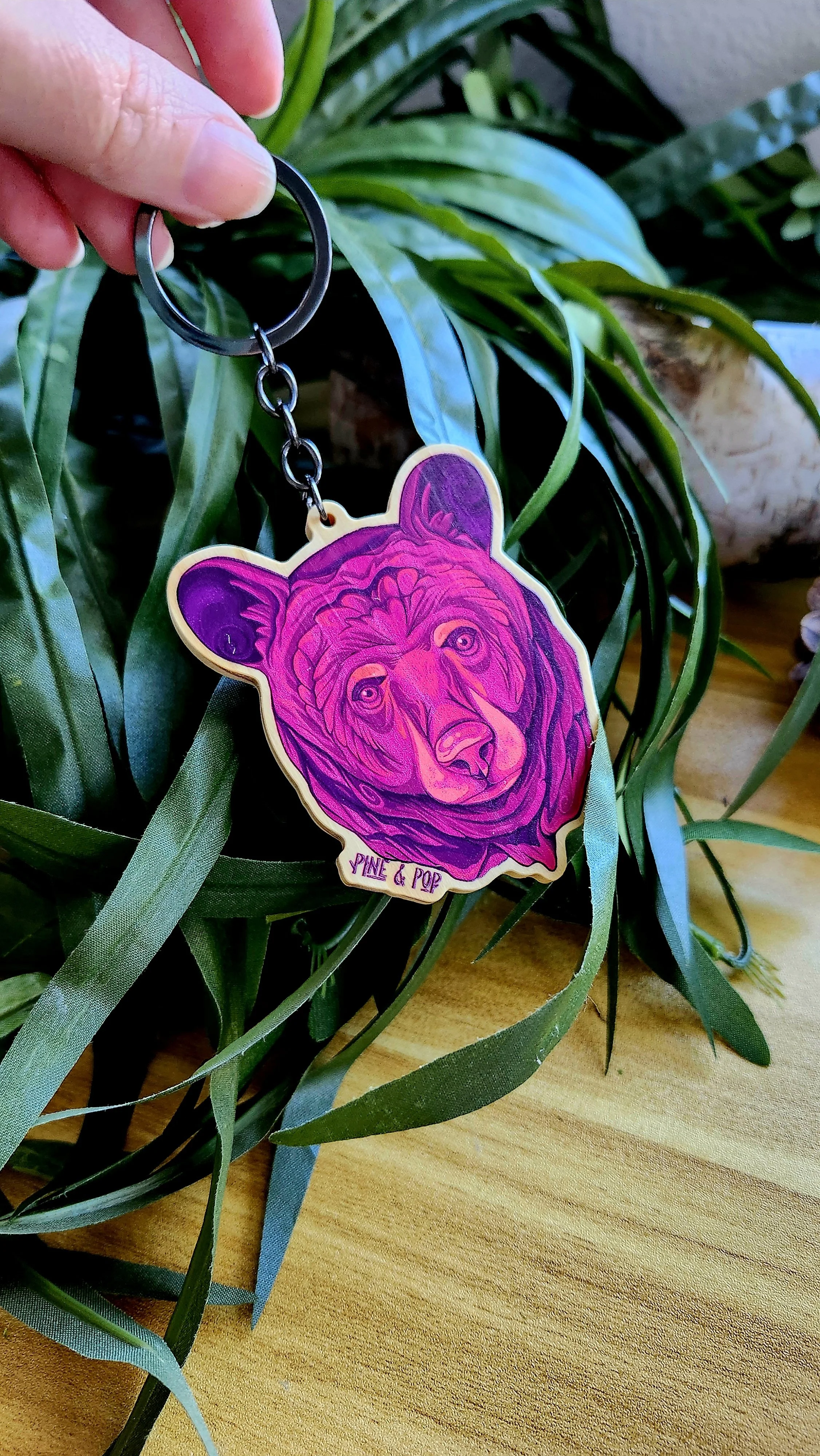 keychain of black bear face with purple, mauve, and peach tones with a gunmetal grey chain and keyring in someone's hand