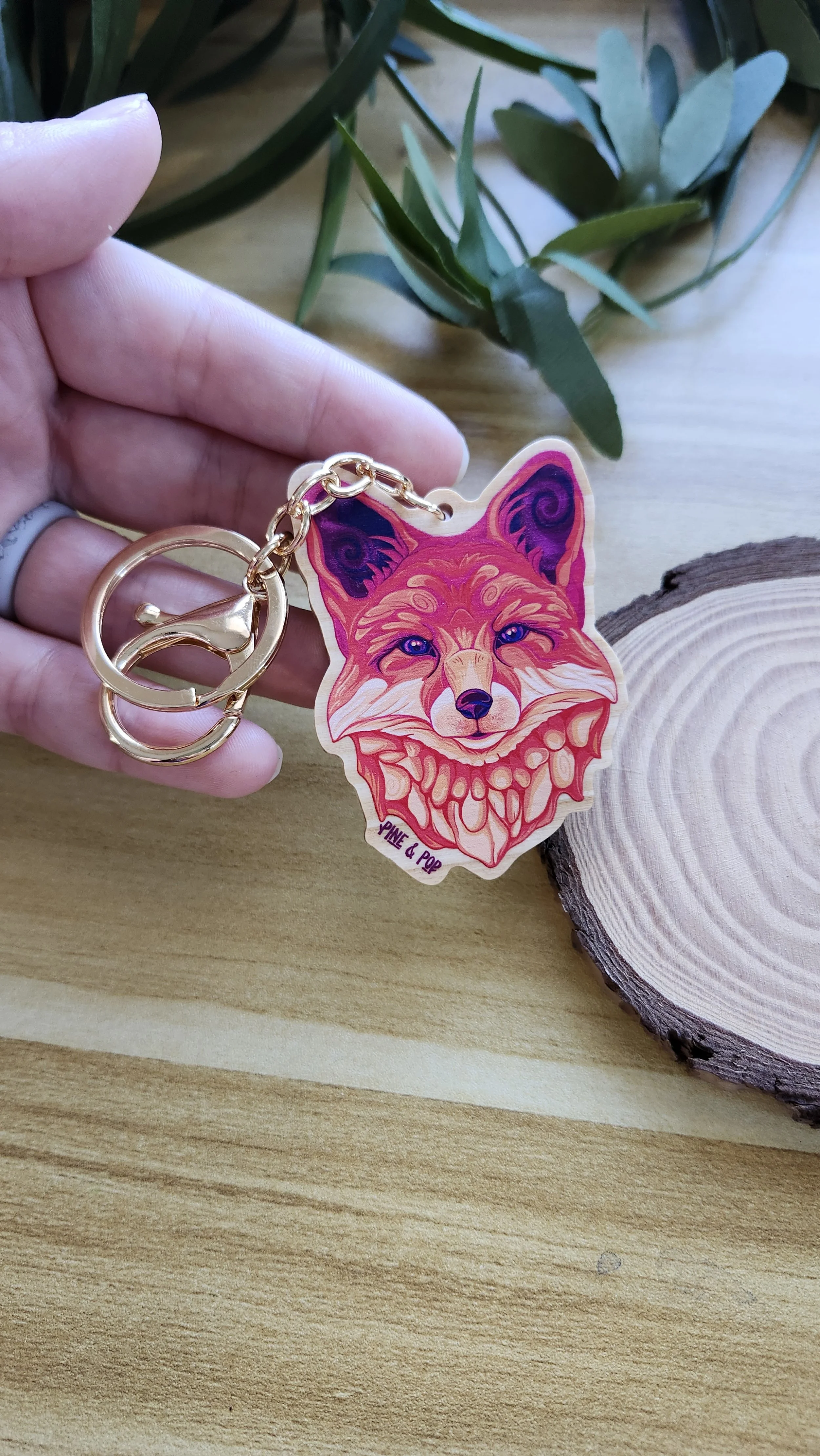 a hand holding a keychain of a fox head with red, orange, cream, and purple colors used to create the design