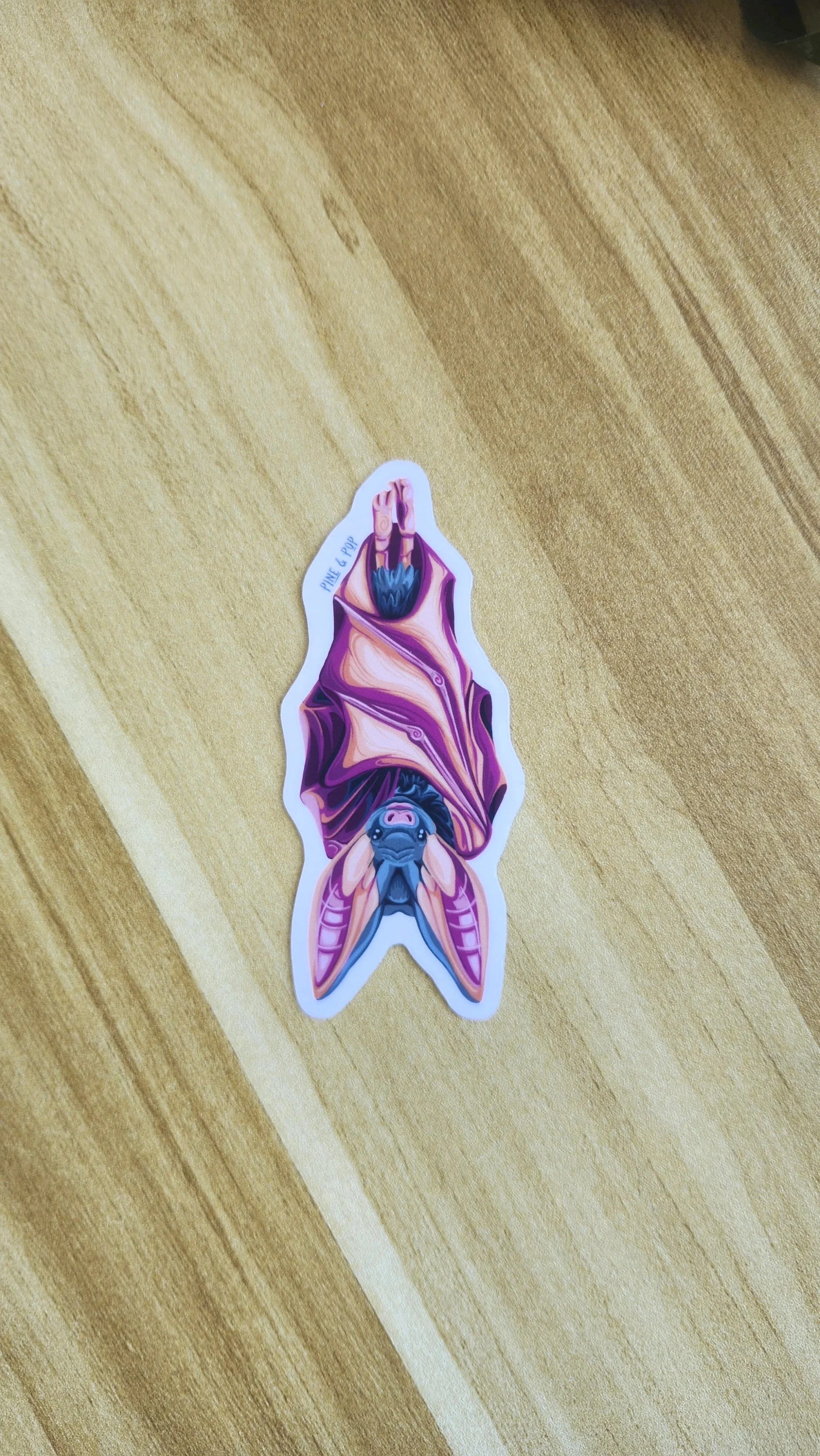 sticker of bat hanging with its wings wrapped around its body designed with orange, teal, pink, and purple colors