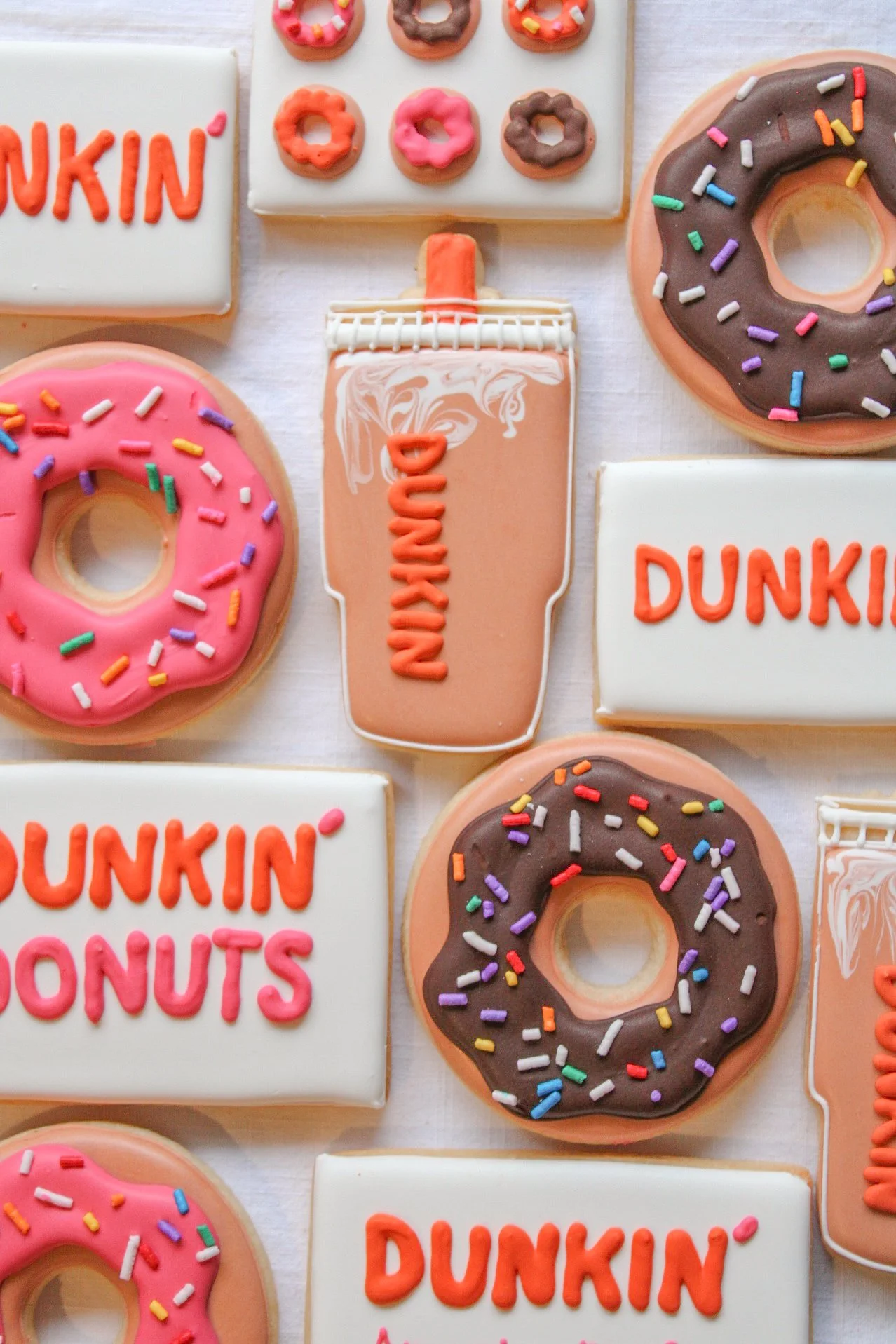 Decorative cookies shaped like donuts, a coffee cup, and rectangular signs with 'Dunkin' and 'Dunkin' Donuts' written on them, decorated with pink, brown, and white icing and colorful sprinkles.