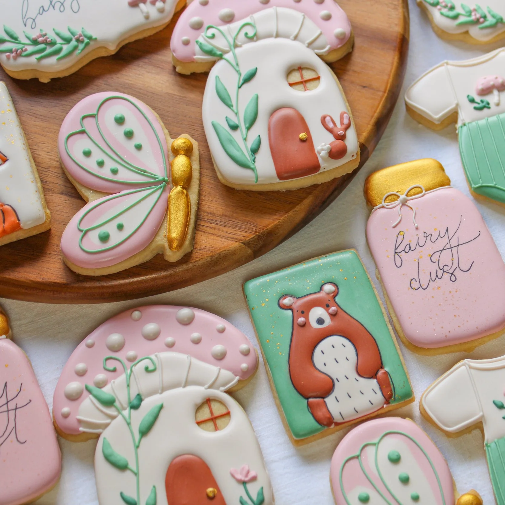 Pastel decorated baby cookies with fairy tale and nature themes, including a butterfly, a bear, a house, and a jar labeled 'fairy dust', arranged on a wooden tray and white surface.