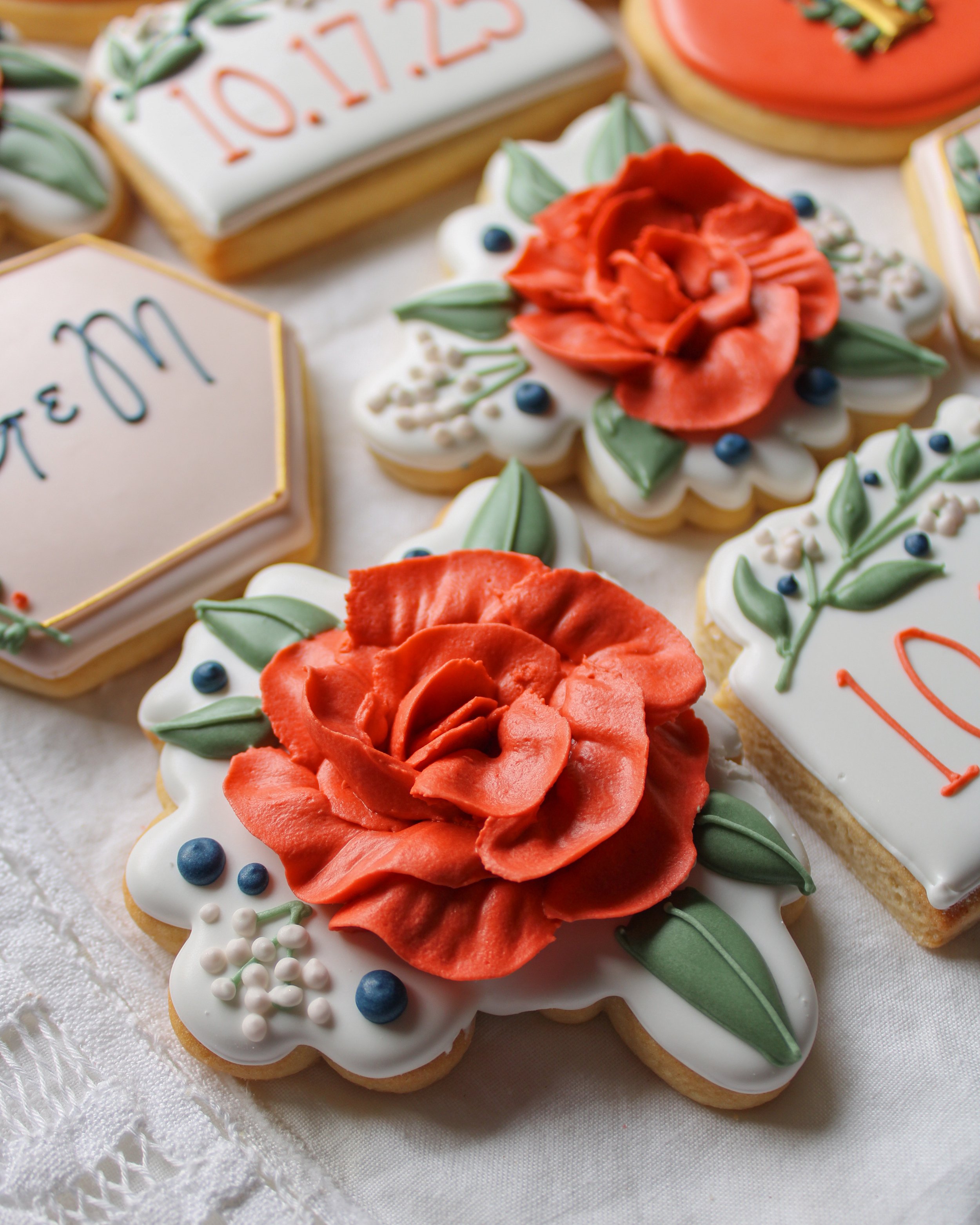 Decorative cookies with red roses, green leaves, and white icing, some with messages written in red and blue icing, on a white tablecloth.