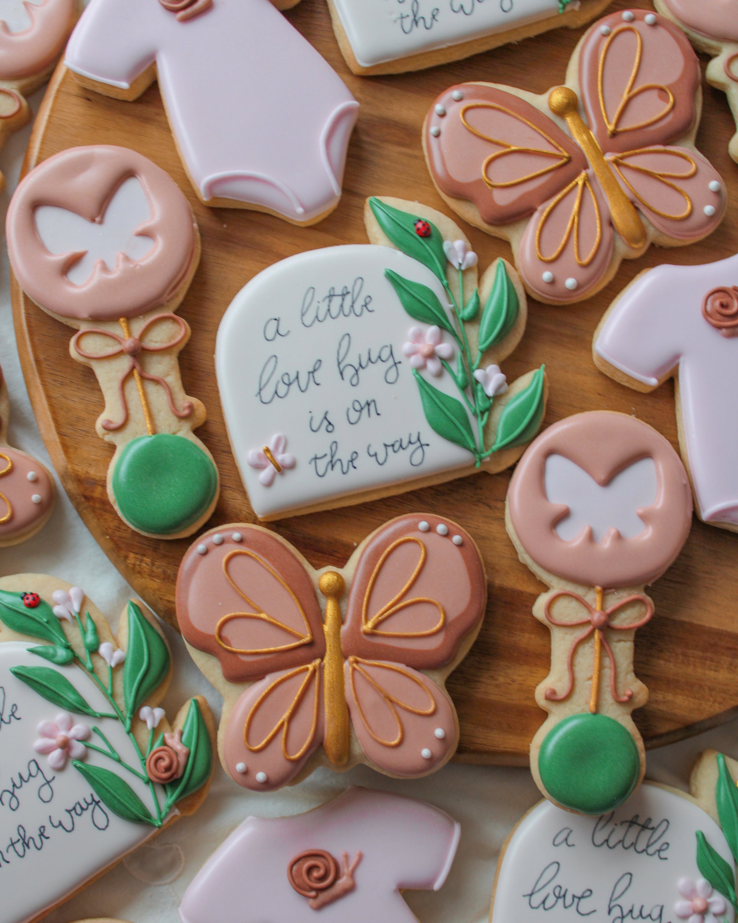 Decorative cookies shaped like butterflies, flowers, and a message reading 'a little love bug is on the way'. The cookies are decorated with pastel-colored icing in pink, white, green, and brown with intricate details.