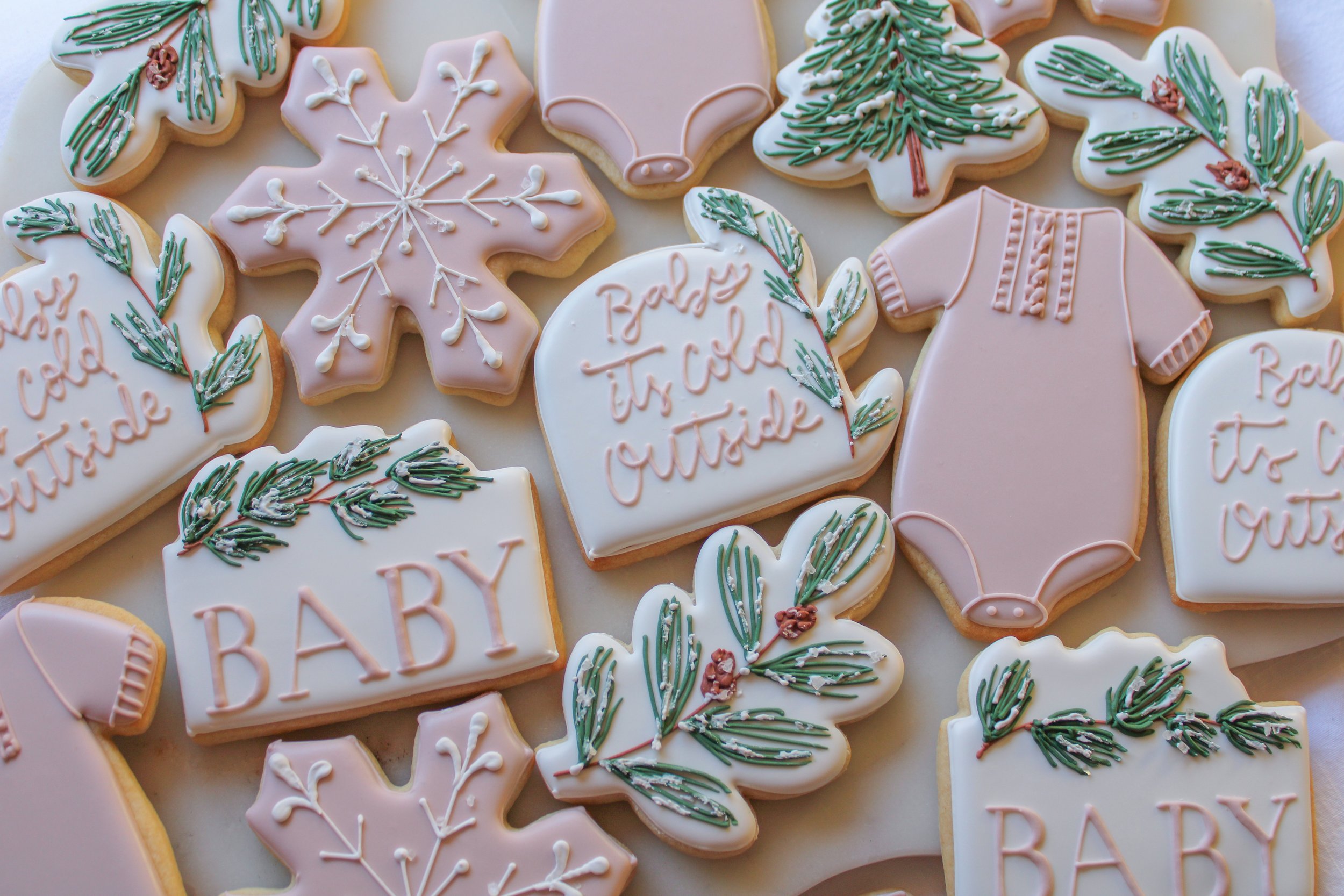 Assorted decorated cookies with baby and nature-themed designs, including pink and white icing, snowflakes, baby onesies, hearts with the phrases 'Baby It’s Cold Outside,' and green foliage accents.