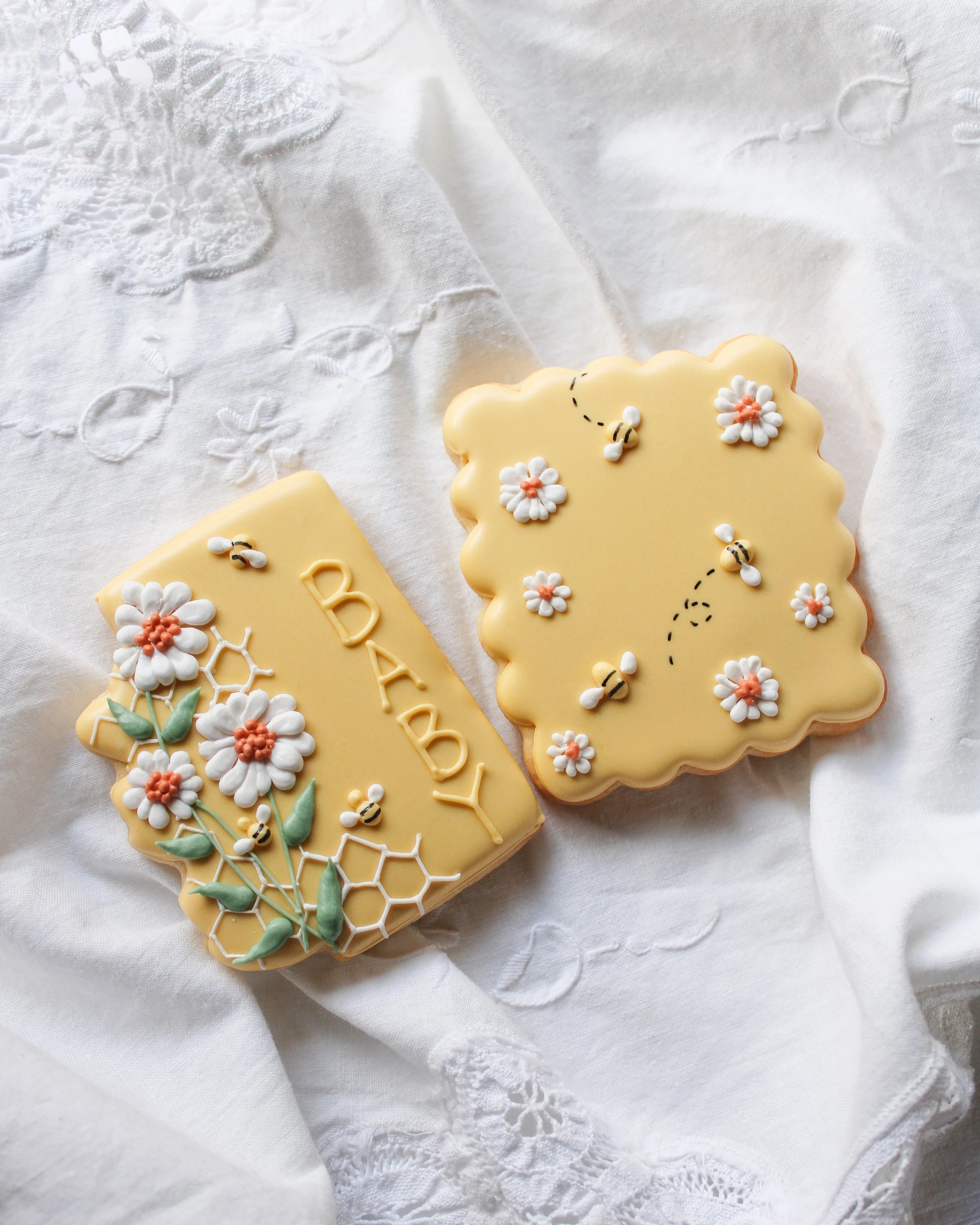 Decorative cookies on white embroidered cloth, one with the word 'BABY' and flowers, the other with bees and flowers.