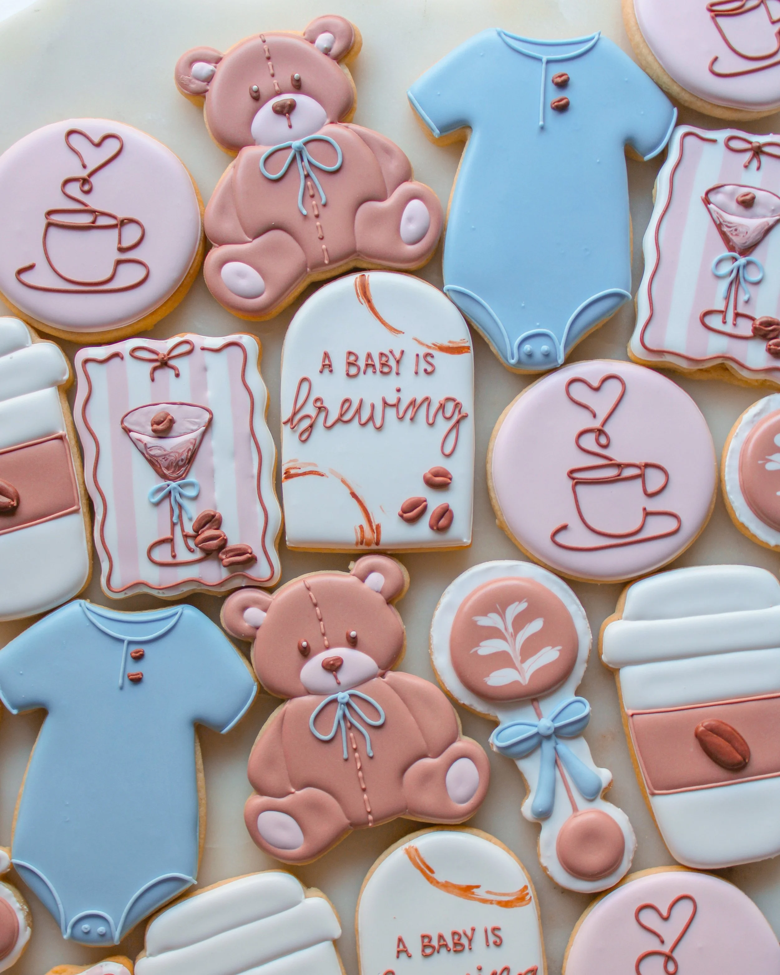 Assorted baby-themed cookies decorated with pastel colors, including teddy bears, baby onesies, bibs, teacups, and a sign that reads "A BABY IS brewing".
