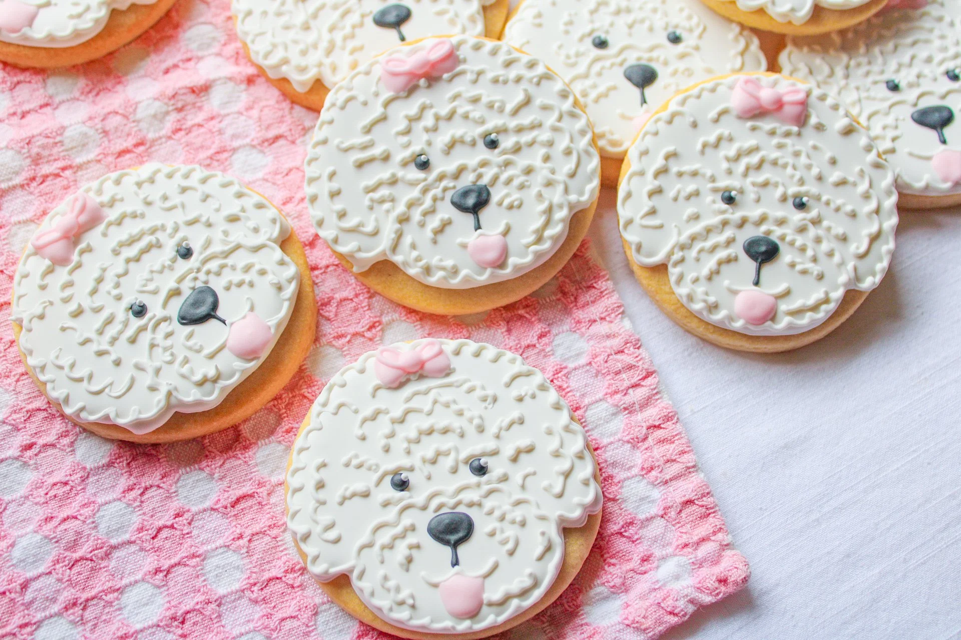 Decorated sugar cookies with a dog's face design, featuring white icing, black icing for the nose and eyes, pink icing for the tongue and bow, on a pink and white polka dot and plain white cloth.