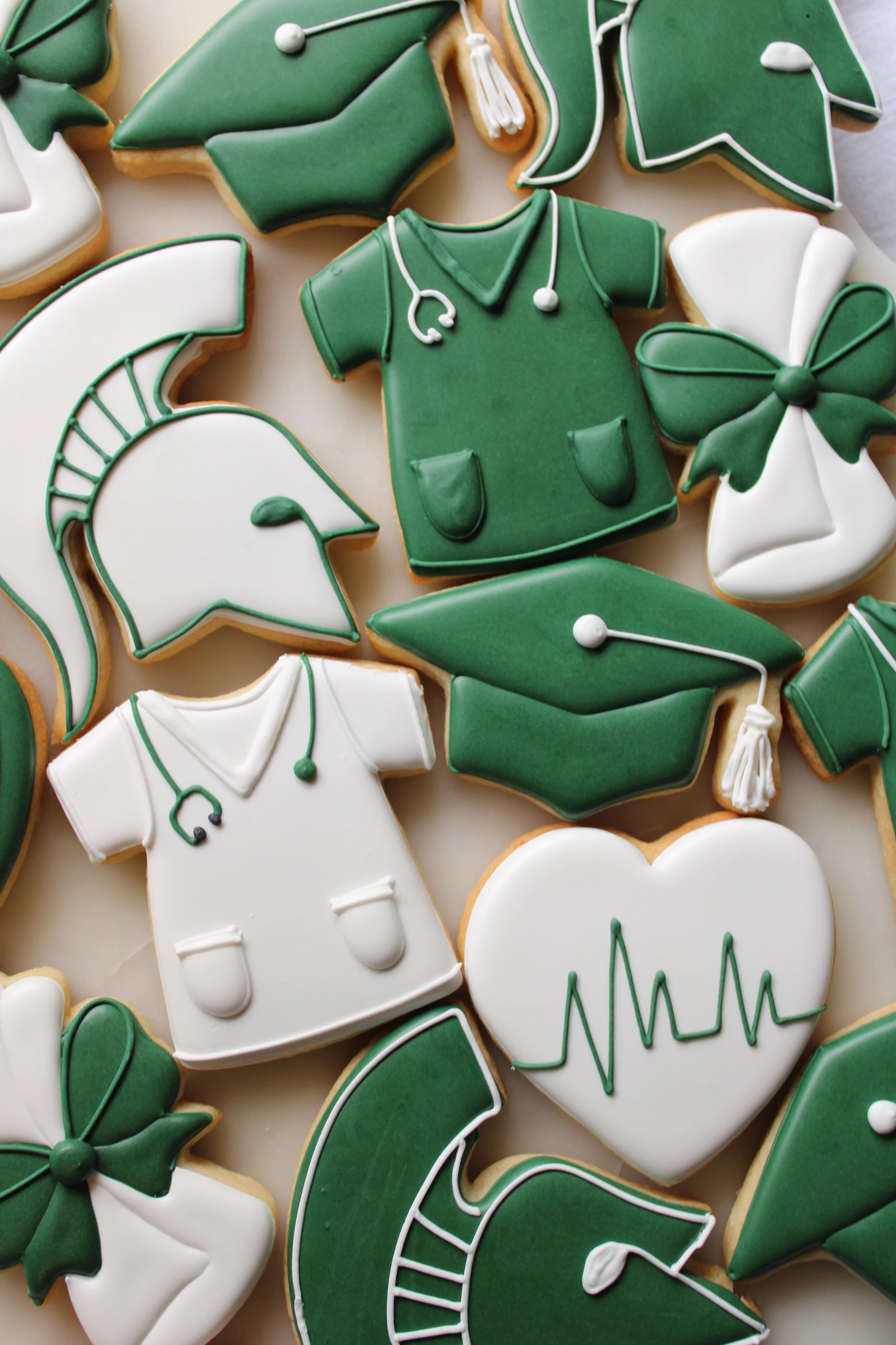 Decorated MSU graduation cookies with medical themes, including stethoscopes, nurse scrubs, graduation caps, and hearts with heartbeat lines, in green, white, and black icing.